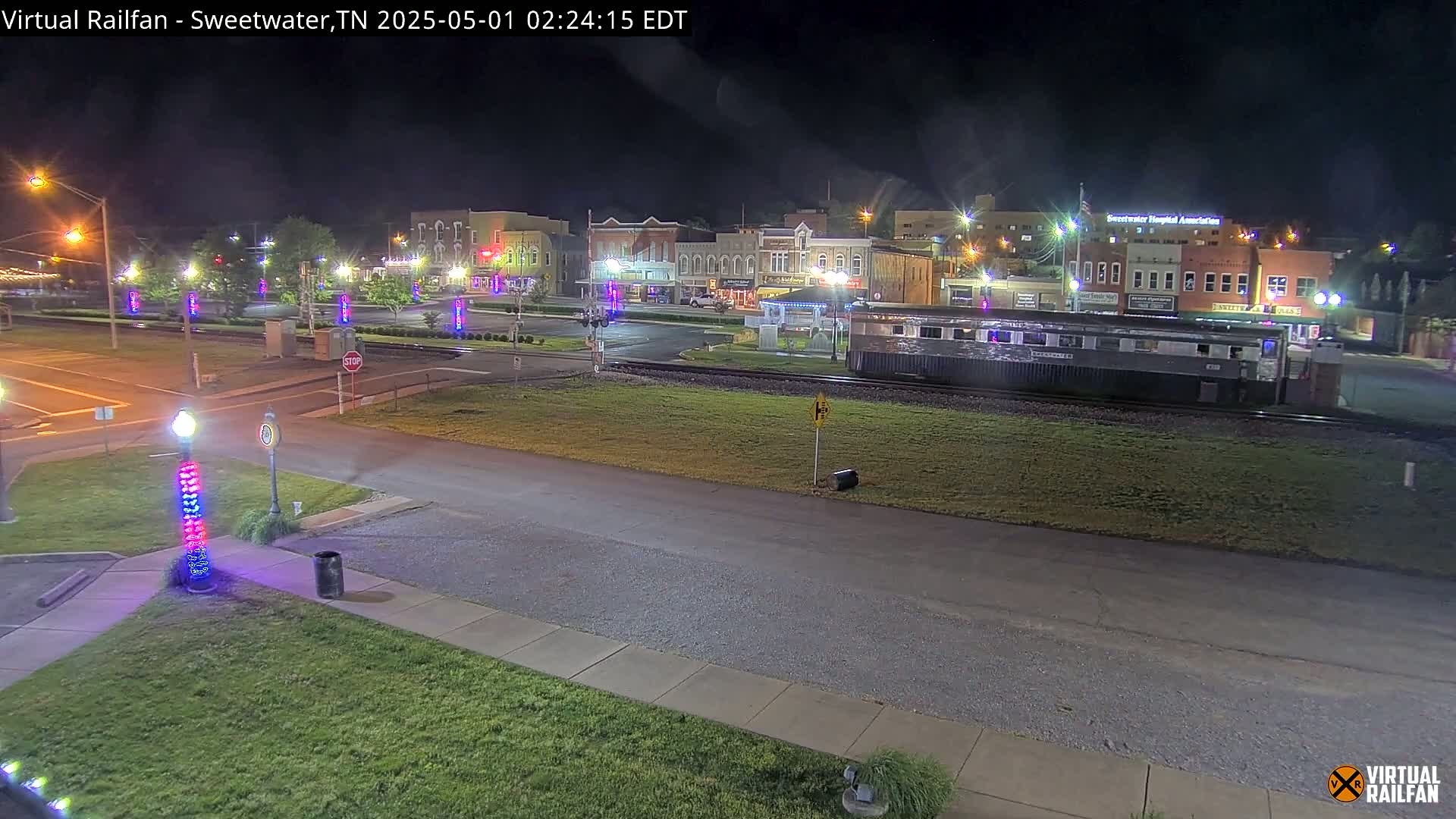 A nighttime view of a town square with a train car parked on the tracks in the foreground, illuminated by streetlights and decorative lights.