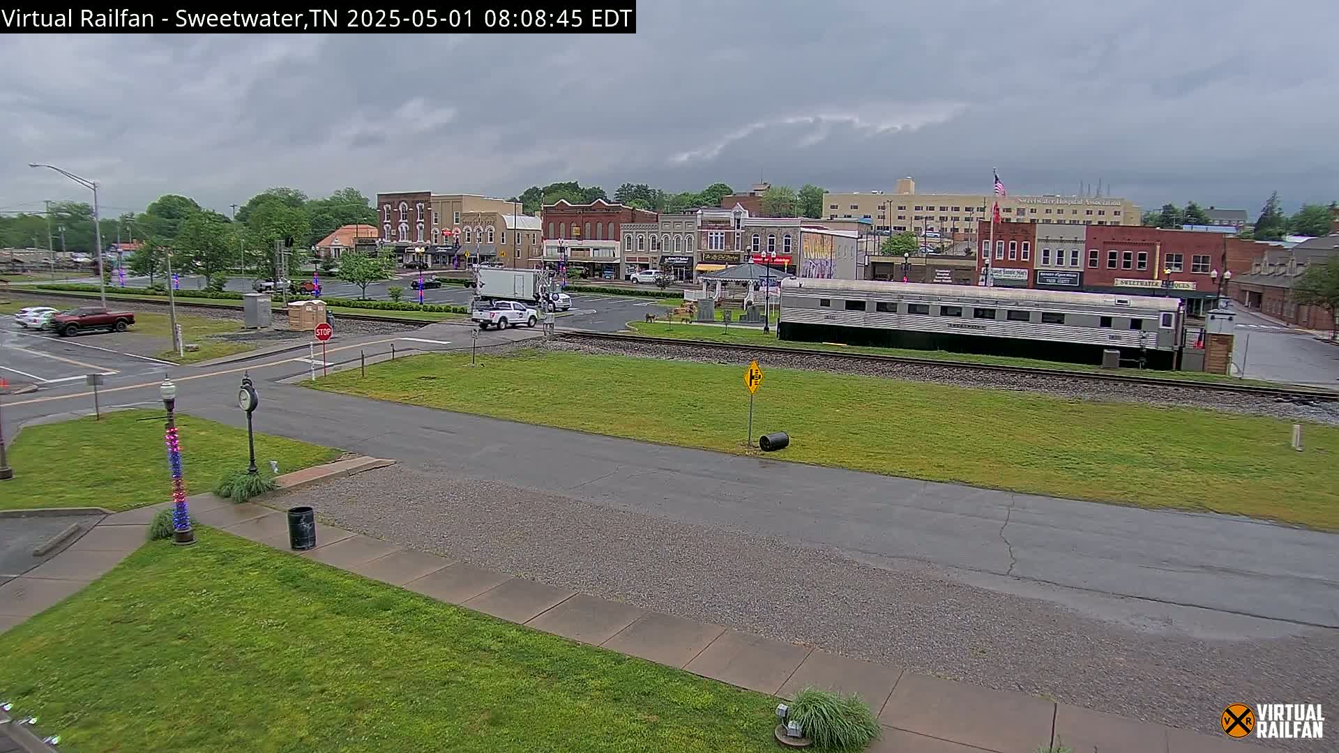 A view of a town square under overcast skies, showing a railroad car parked on tracks near several brick buildings and a grassy area.