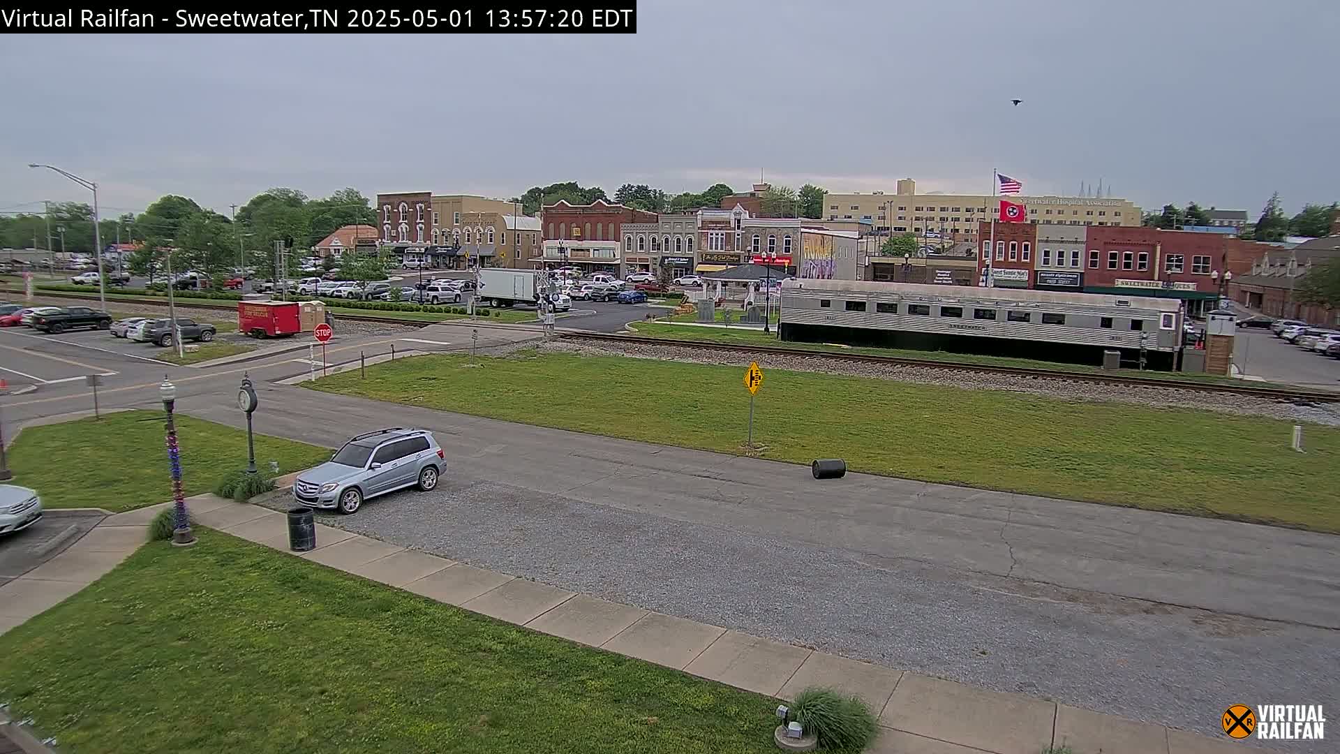 A partly cloudy day reveals a small town's streetscape, featuring parked cars, a railroad track with a passenger car, and several buildings in the background.