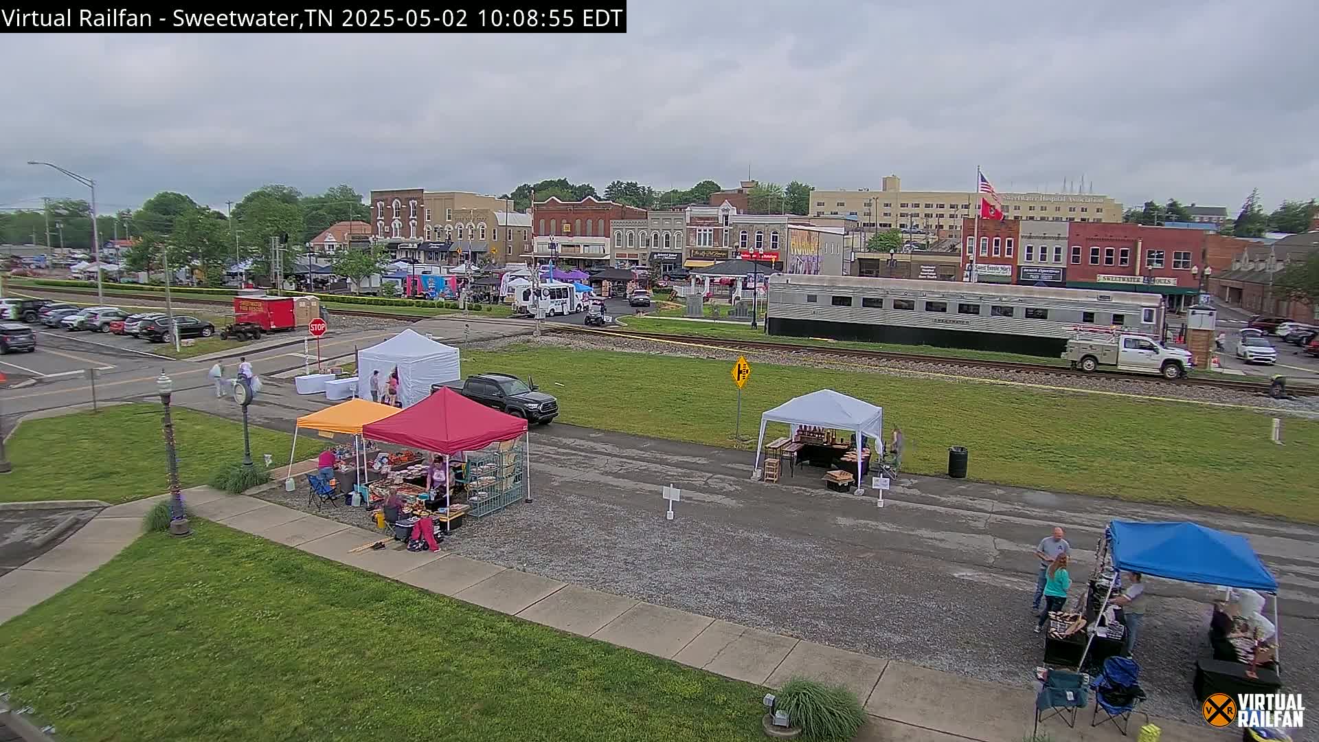 An outdoor market with various vendors under tents is set up near a railroad track and in front of a town's buildings under an overcast sky.