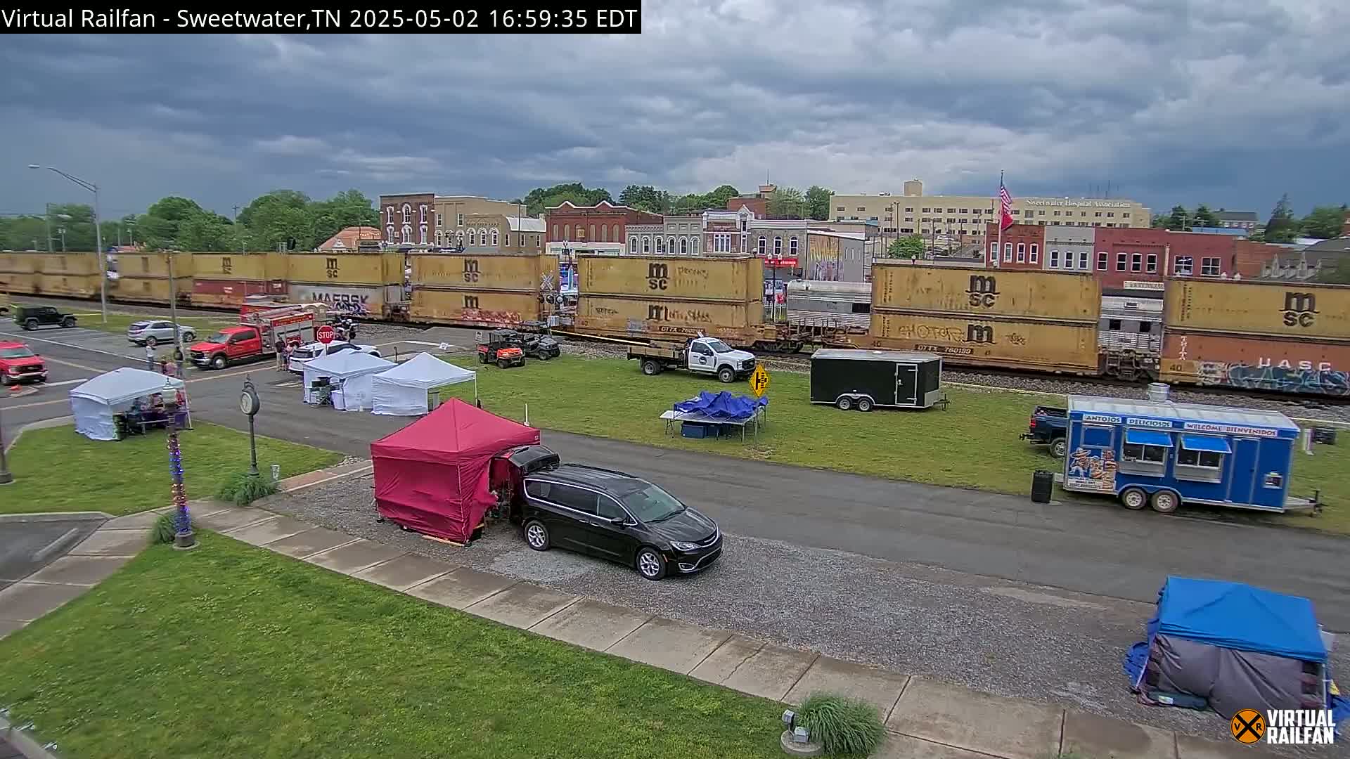 A long freight train passes through a town square under a cloudy sky, where several tents and food trucks are set up.