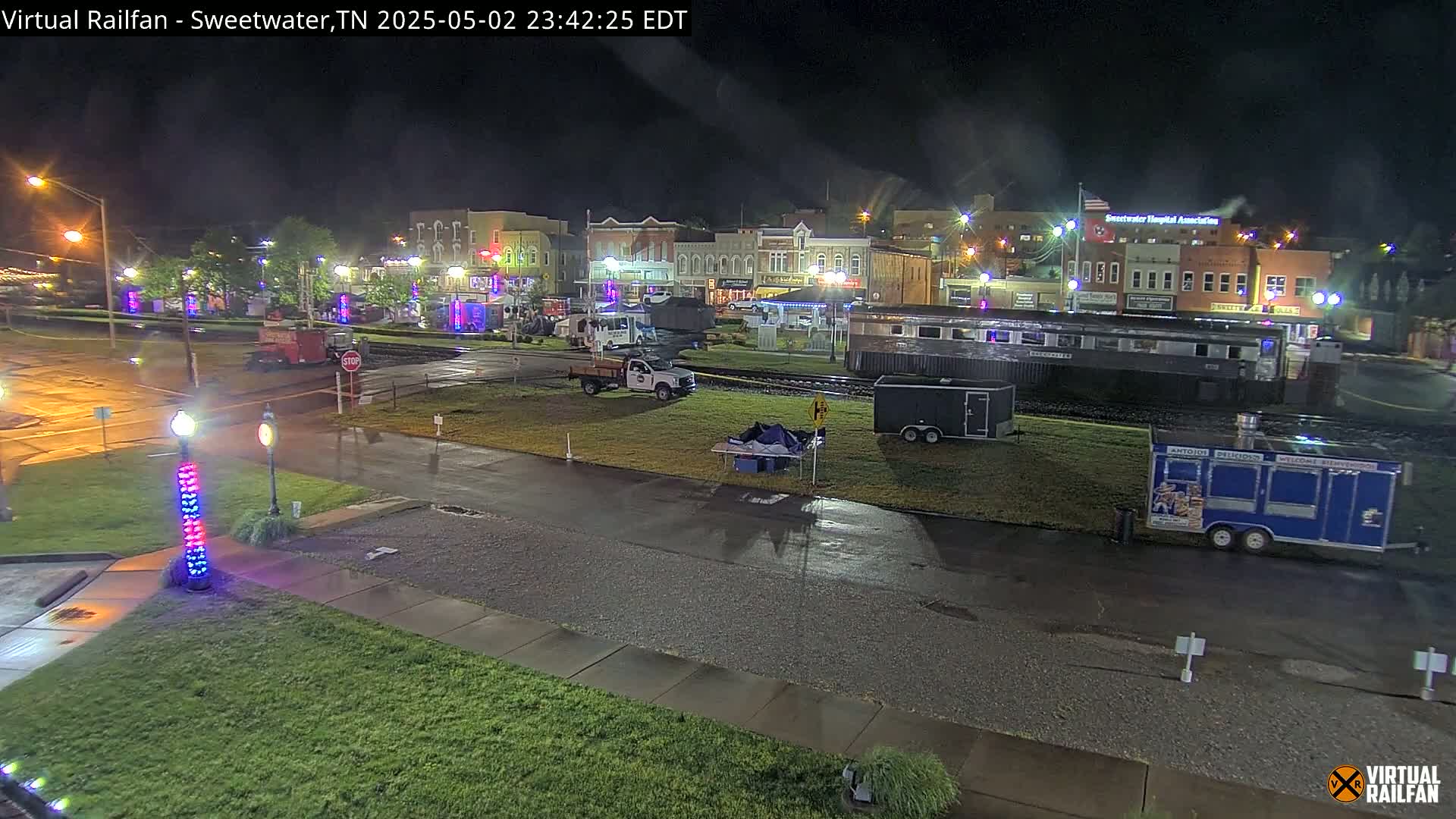 A nighttime view of a town square with several vehicles parked near a railway car and food trucks, under wet conditions.