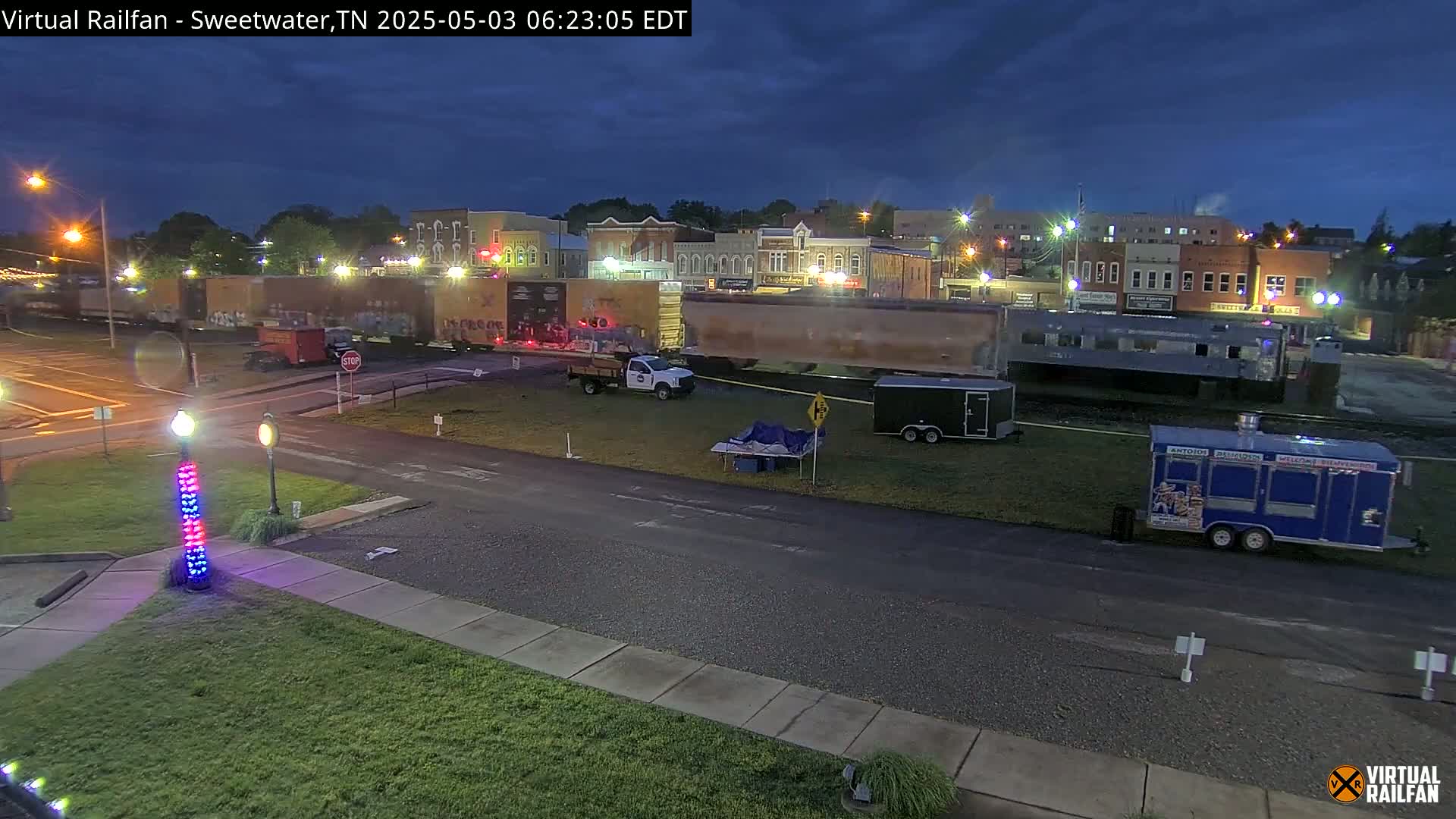 Under a dark, partly cloudy night sky, several freight cars are parked near a town's illuminated buildings, with a few vehicles and a food truck nearby.