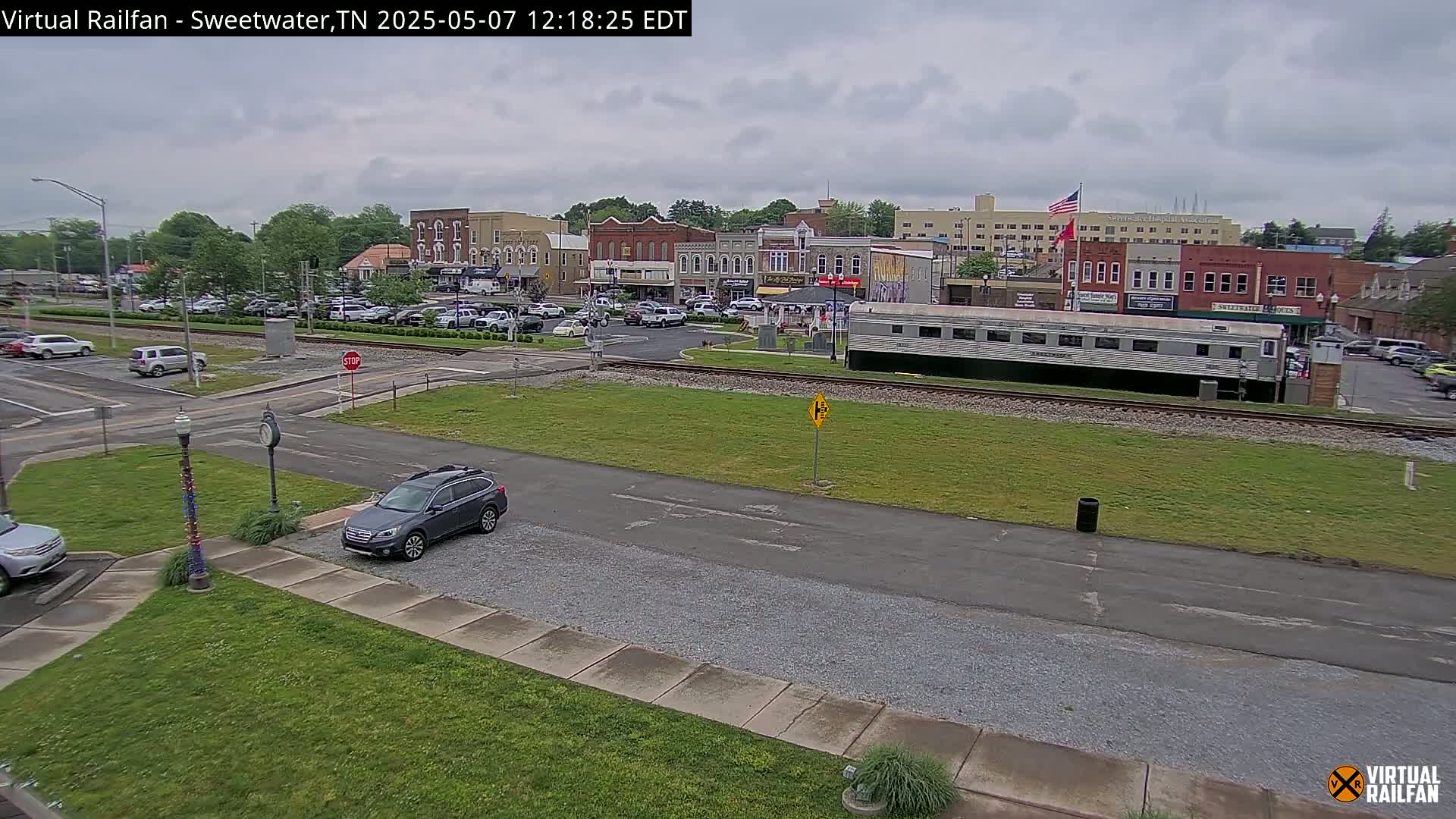 A gray SUV is parked near a grassy area next to railroad tracks, behind which sits a town under an overcast sky.