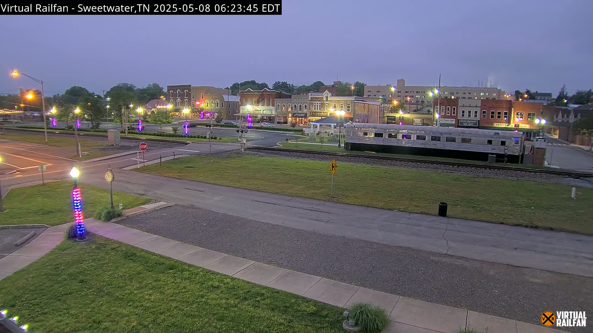 A long, silver train car sits on railway tracks in front of a town square at dusk, with several buildings visible under a twilight sky.