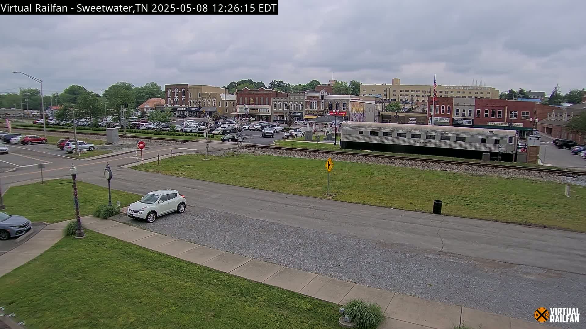 A small town square with overcast skies, a railroad track running through it, and a silver passenger car parked next to the tracks, with cars parked in a lot across the street from the tracks.