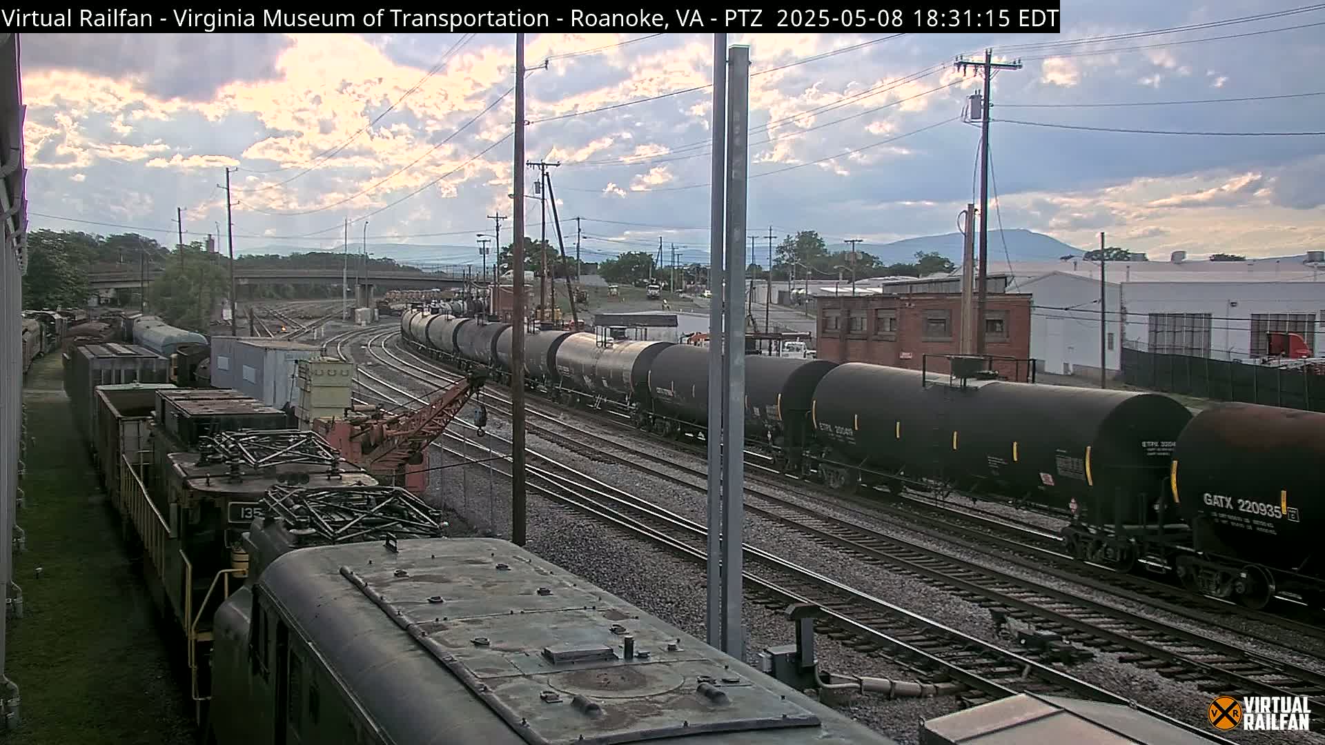 Several freight trains, including tank cars, are visible on a railway under a partly cloudy evening sky.