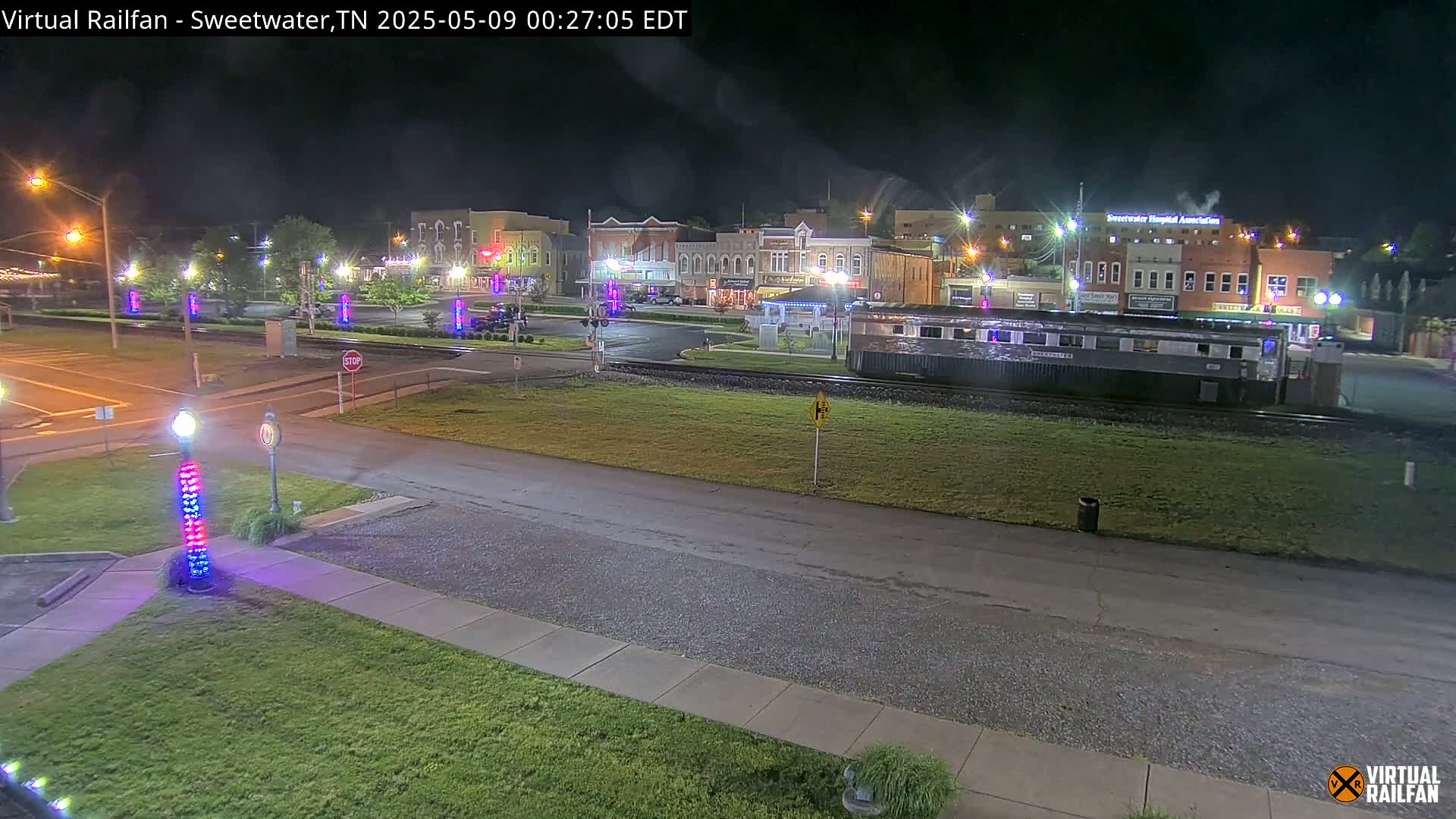 A nighttime view of a town square with a train car parked on tracks in the foreground, under clear skies.