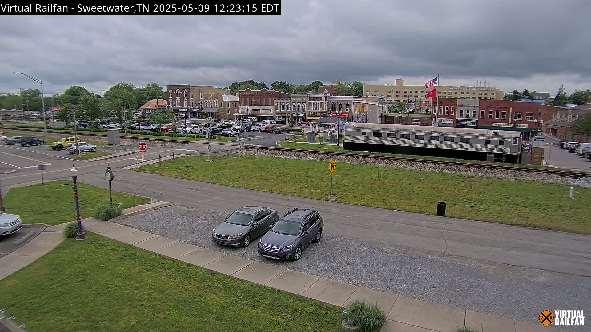 A small town square with overcast skies, featuring several parked cars, a long silver train car, and multiple brick buildings.