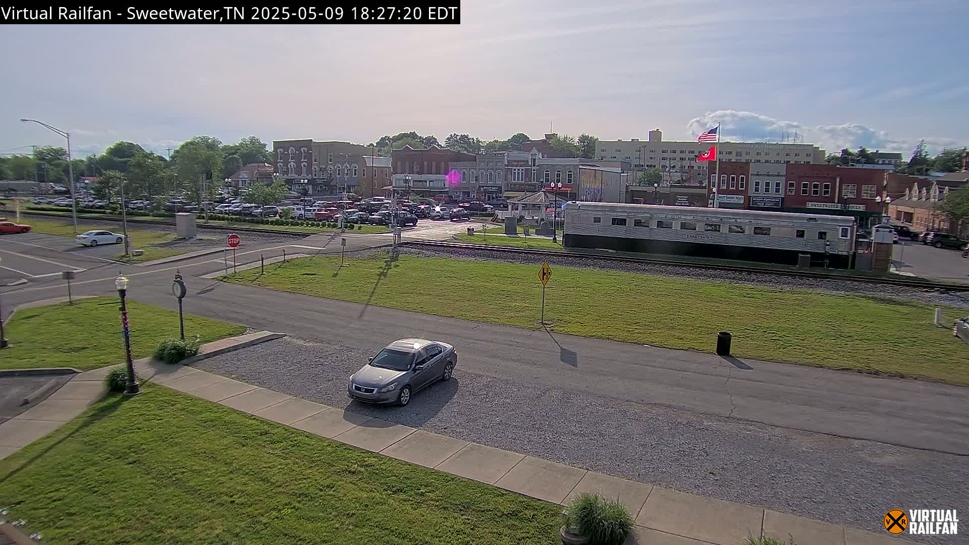 A gray car is parked in a lot next to a railroad track where a long silver train car is stopped, across from a town square filled with cars under a partly cloudy sky.