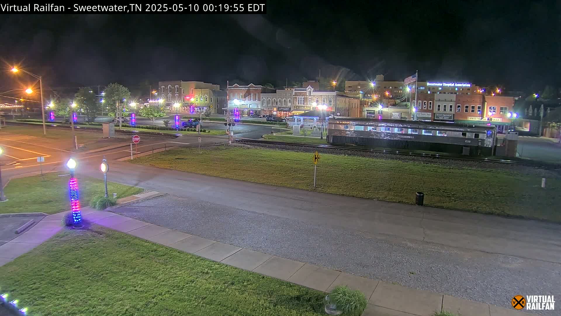 A nighttime view of a town square with a train car parked on tracks in the foreground, under clear skies.