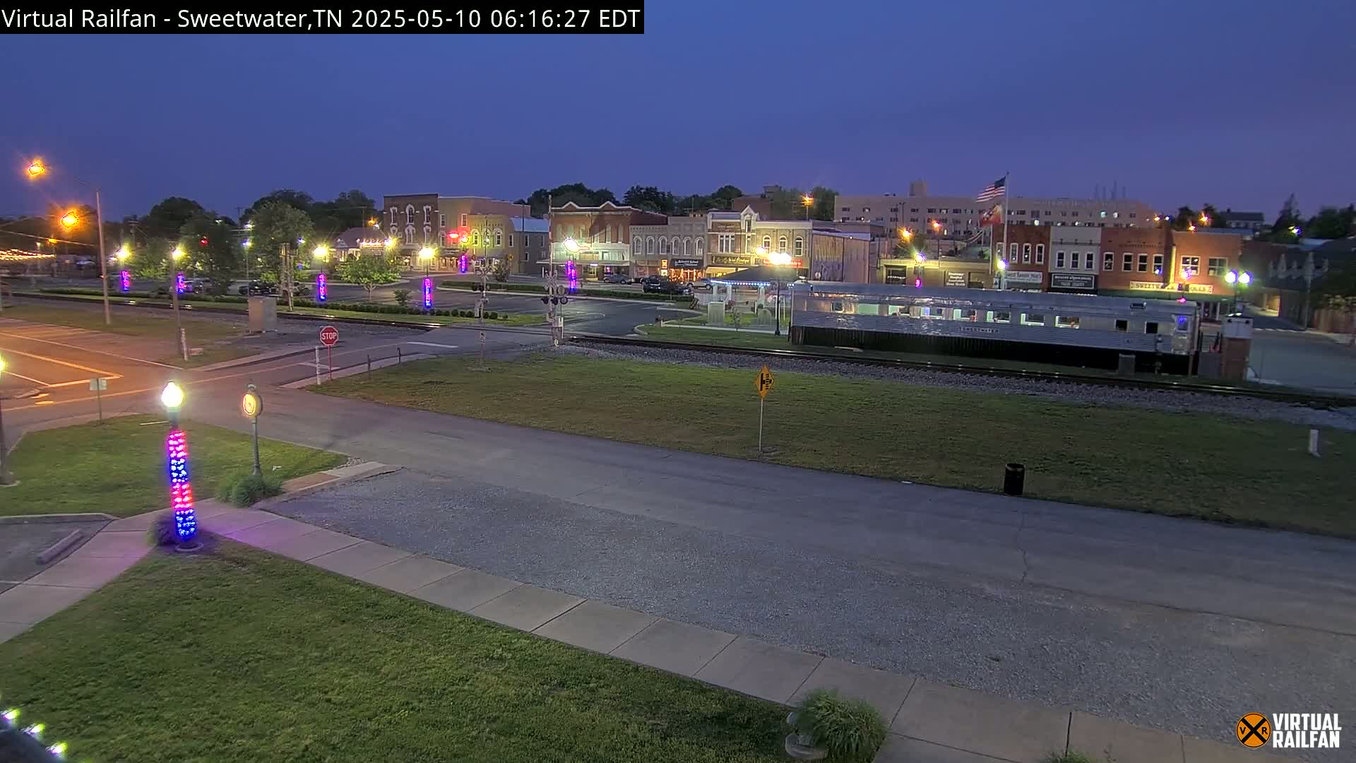 A long, silver train car sits on train tracks in front of a town square at dusk, under a twilight sky.