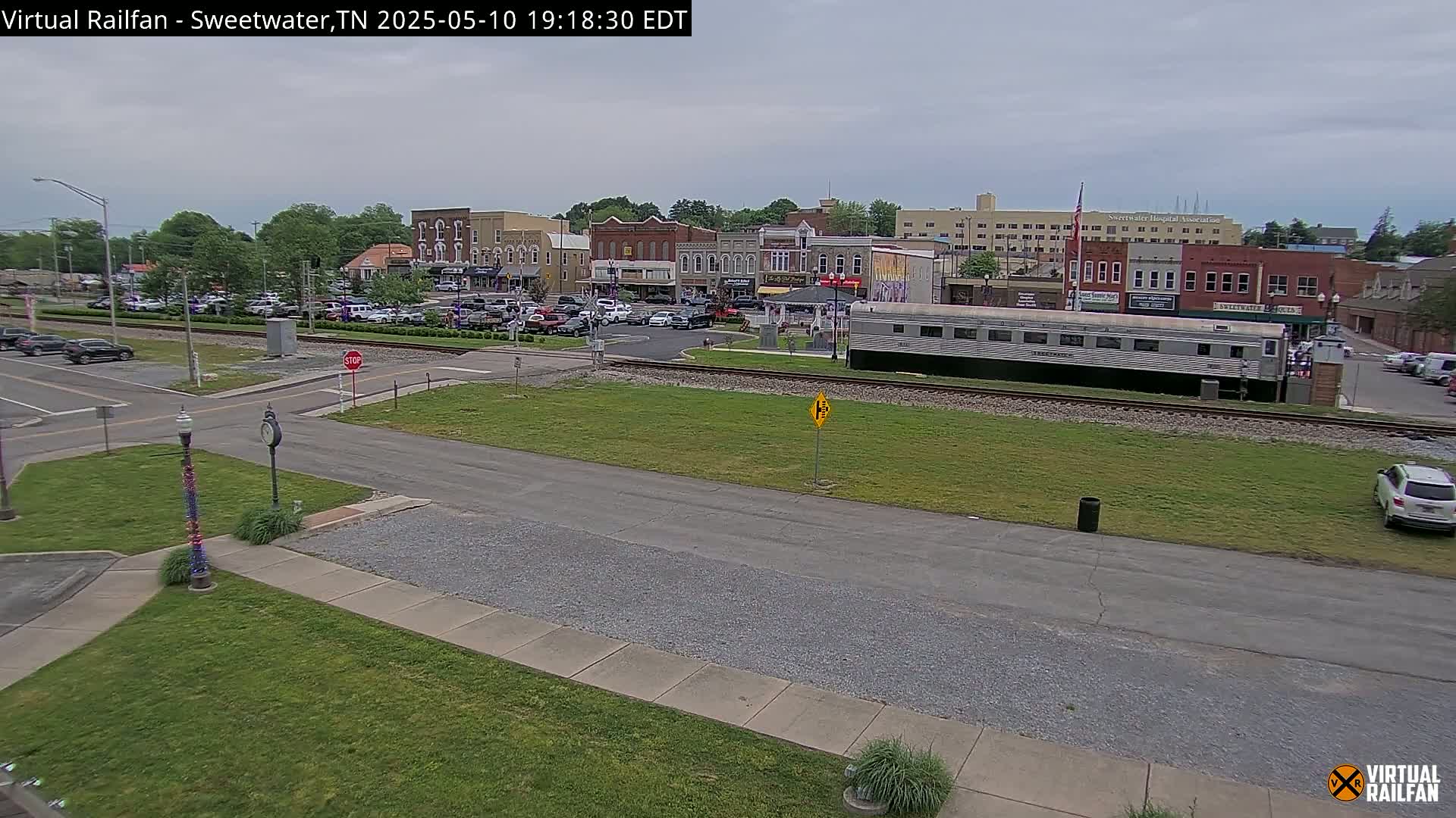A mostly cloudy day reveals a small town square with parked cars, a train car on the tracks, and a few cars on the roads nearby.