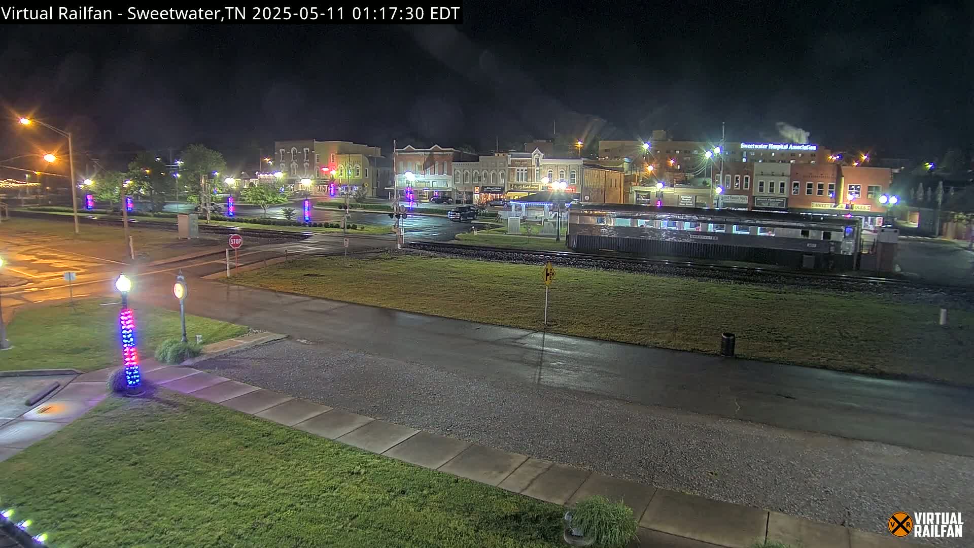 A nighttime view of a town square with several buildings, a railway track featuring a passenger car, and wet streets under partly cloudy skies.