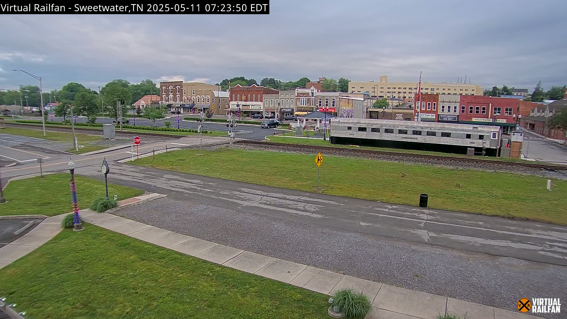 A view of a small town's downtown area on an overcast day, featuring several brick buildings, a railway crossing with a long silver train car, and green lawns.