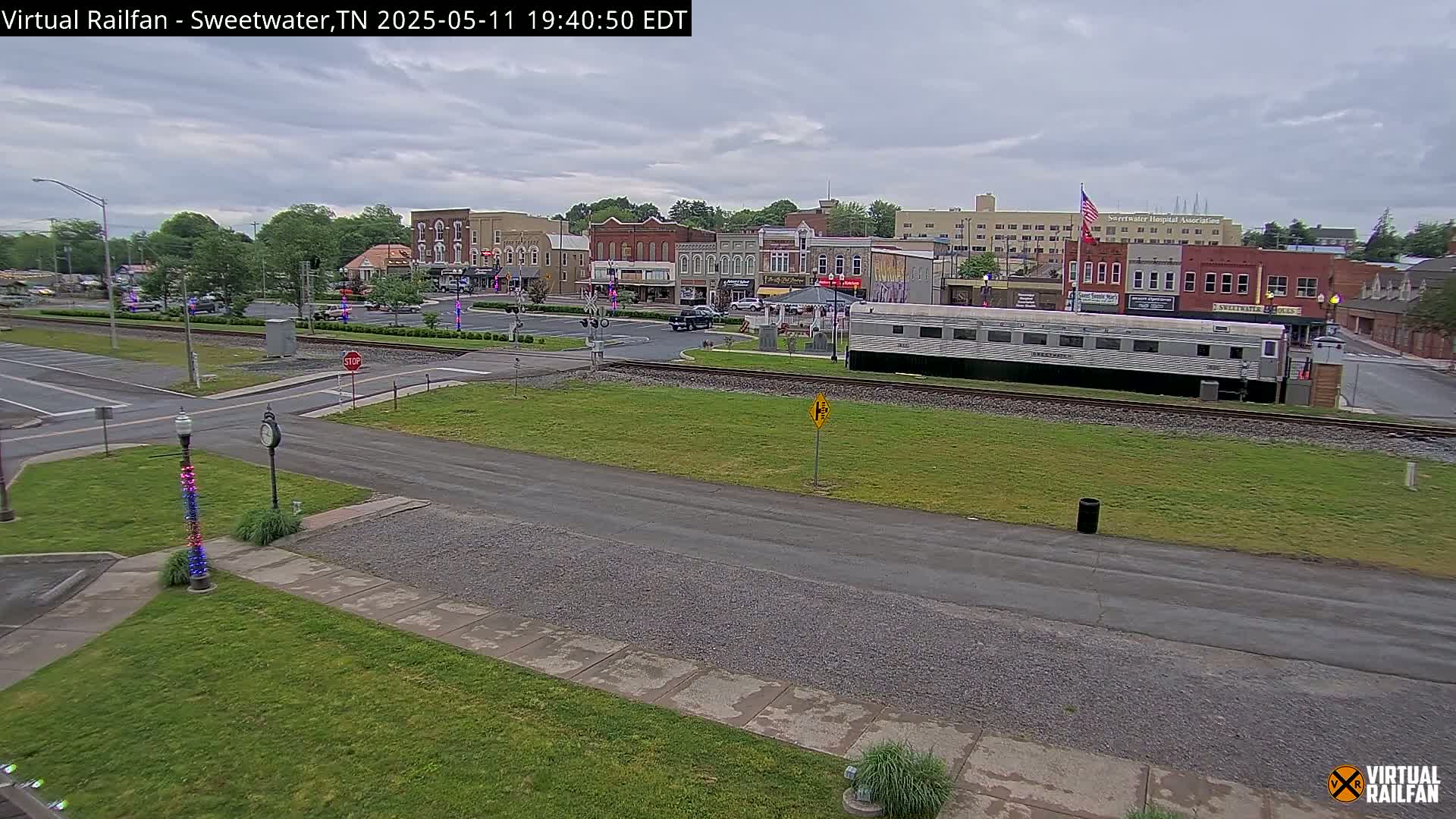 A mostly cloudy daytime scene shows a town square with brick buildings, a railway with a long silver passenger car, and a grassy area with a roadway.
