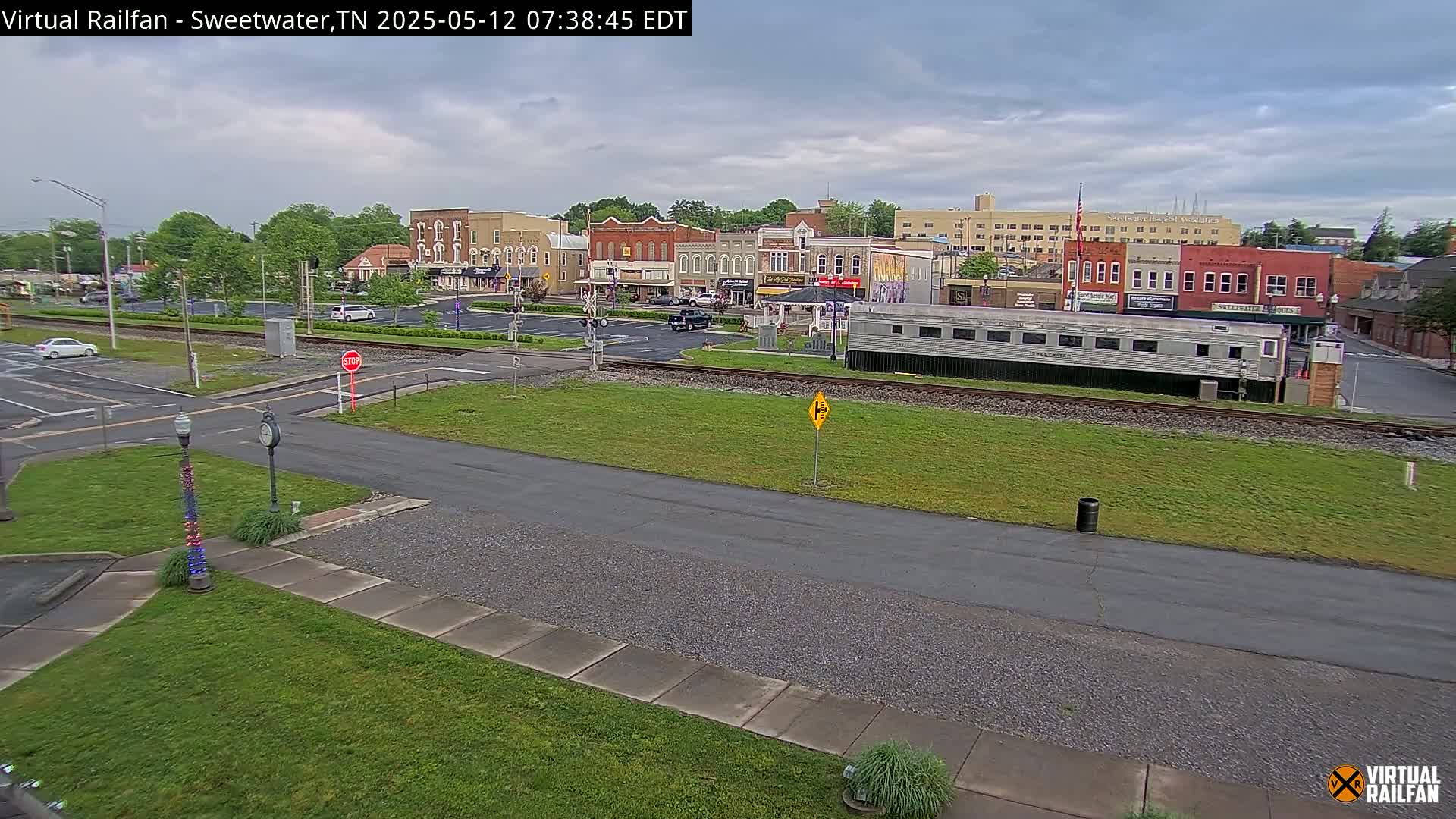 A mostly cloudy day reveals a small town's downtown area, featuring several buildings, a railway crossing, and a long silver passenger car parked near the tracks.