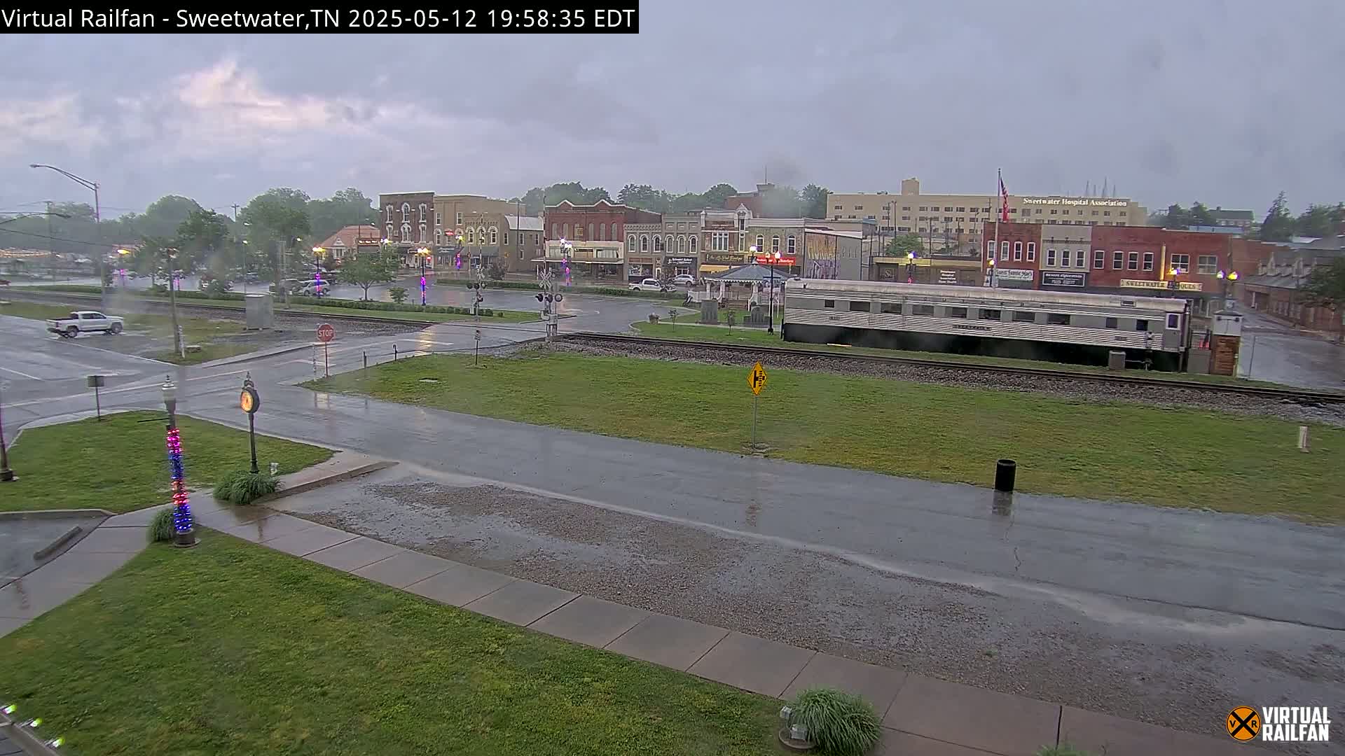 A rain-slicked town square with a silver passenger train car and several buildings visible under a cloudy sky.