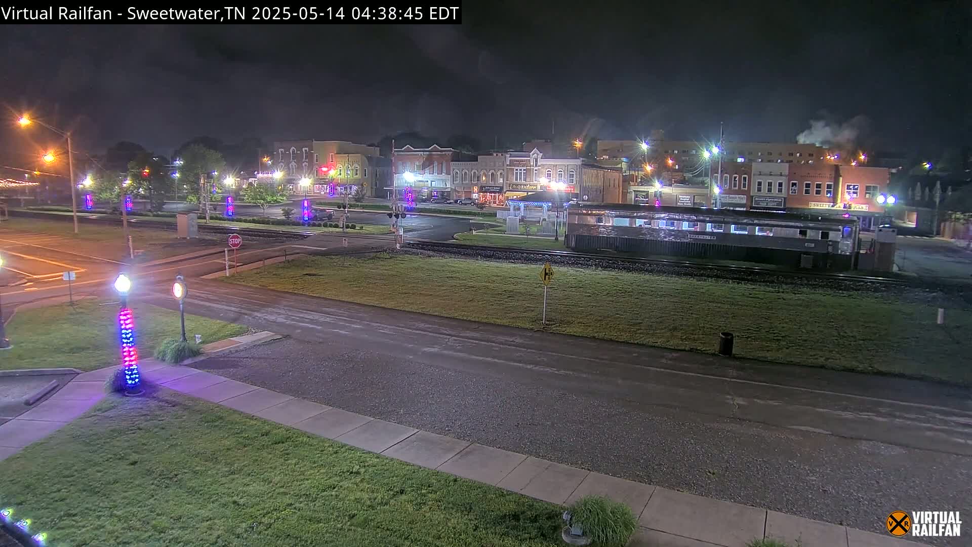A nighttime view of a small town square with a railroad car parked nearby, under partly cloudy skies.