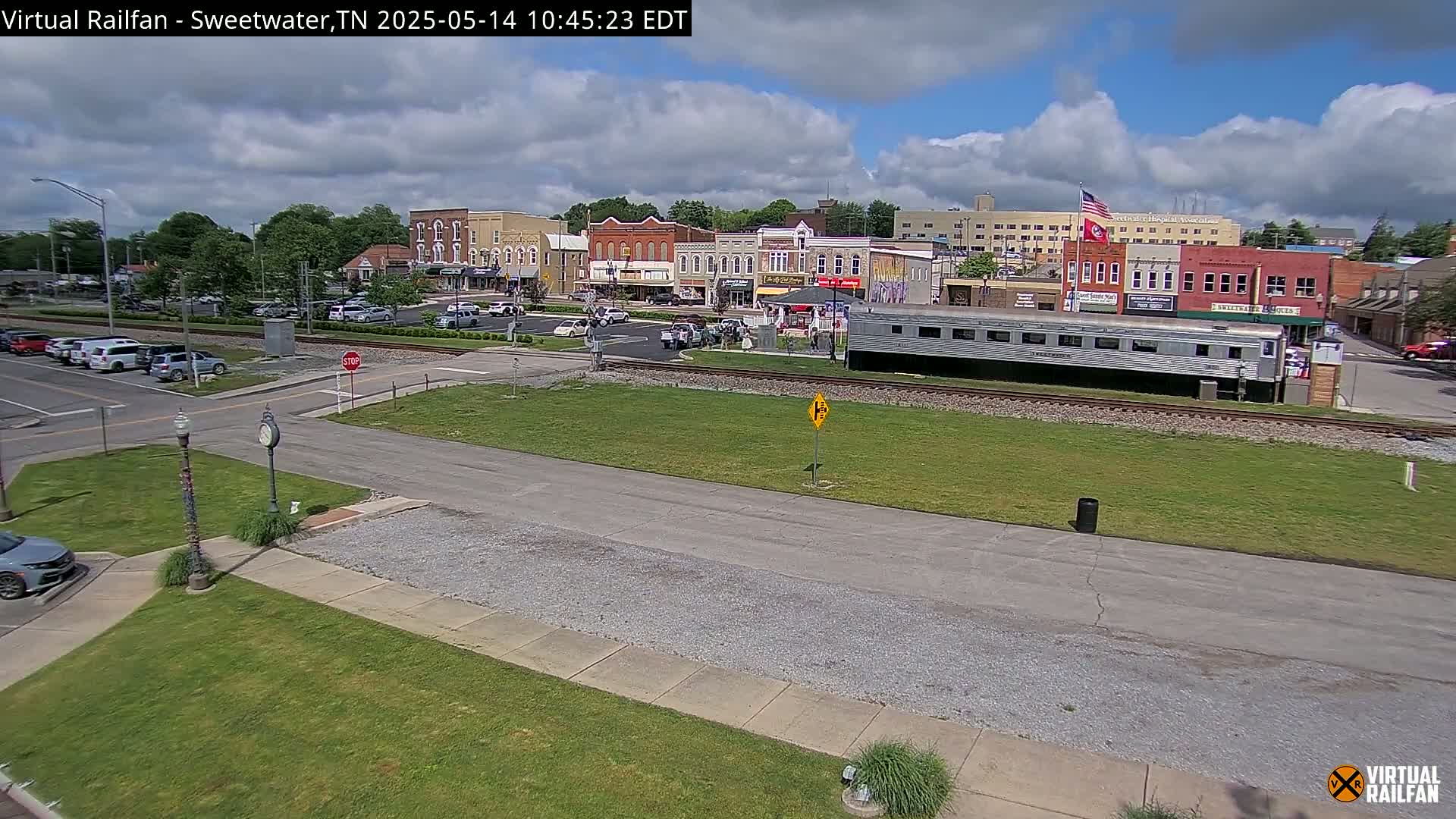 A partly cloudy day shows a small town's downtown area with a railroad track running through it, featuring several buildings and a parked railway car.