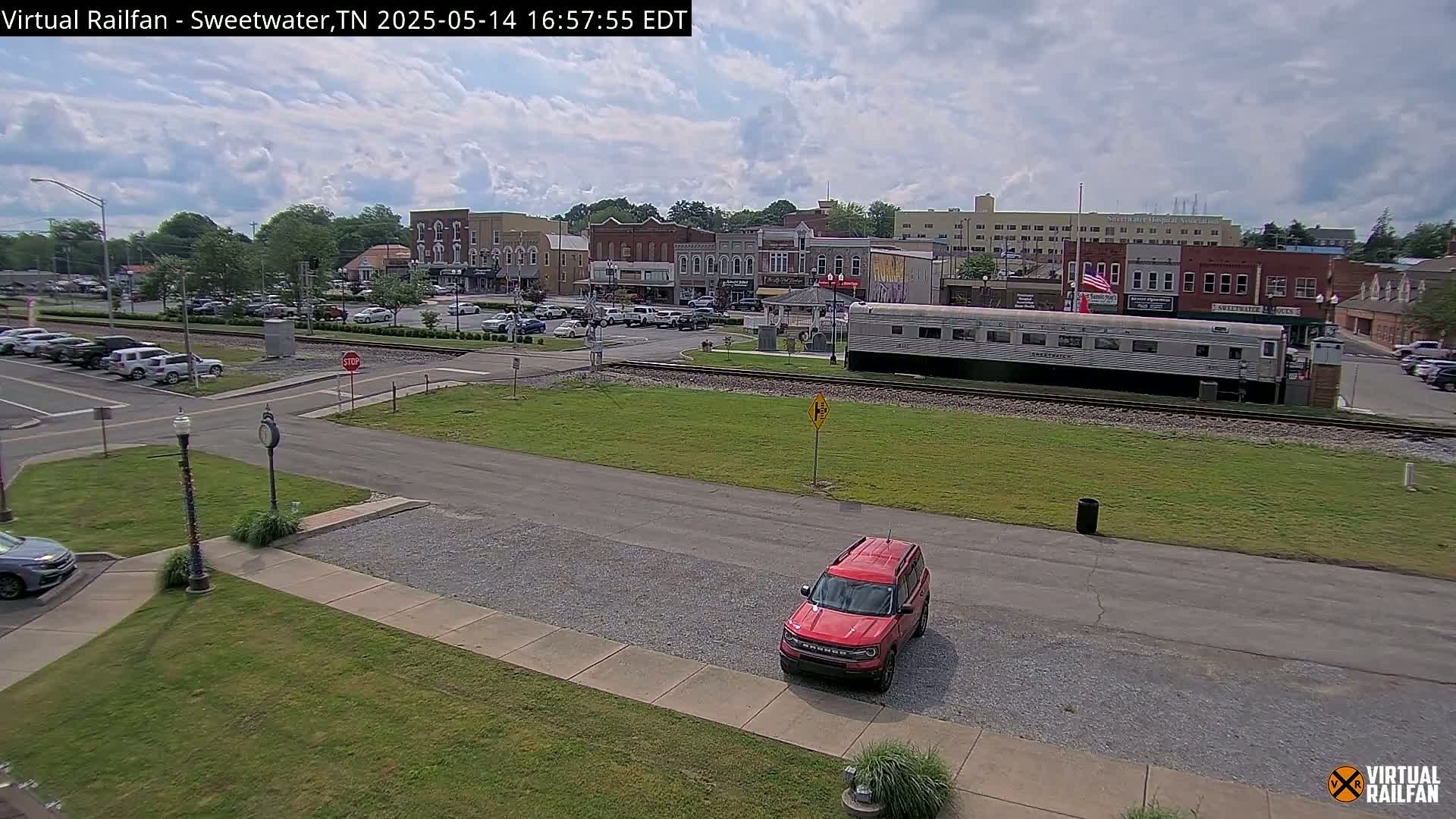 A partly cloudy day shows a small town square with parked cars, a red SUV, and a long silver train car sitting on the tracks.