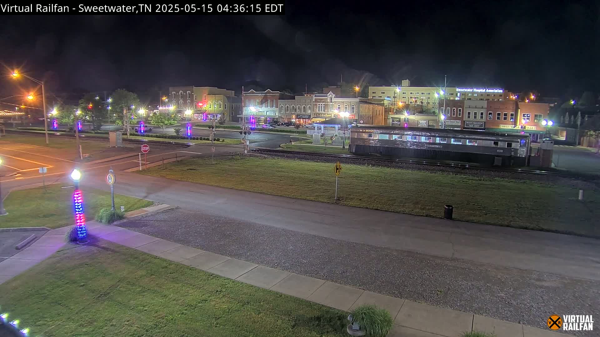 A nighttime view of a town square with buildings illuminated, a railway car on the tracks, and decorative lights along the streets.