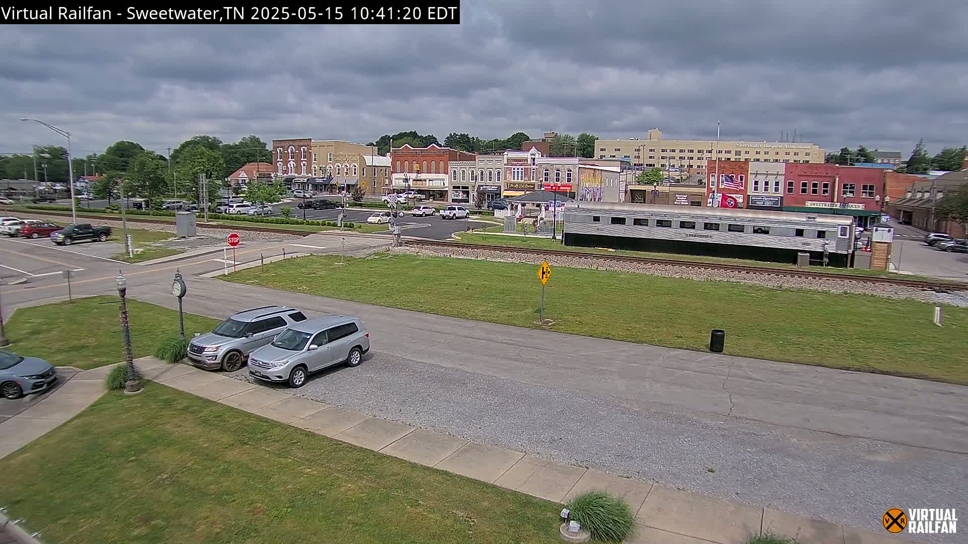 A mostly cloudy day shows a small town's downtown area with several parked cars, a railway, and a silver passenger car, all situated near a grassy area.
