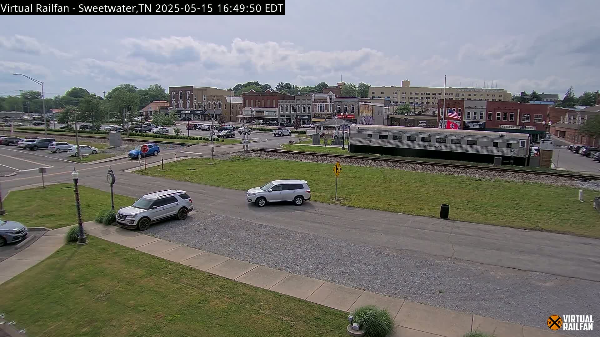 A partly cloudy day shows a town square with several cars, two SUVs, and a long silver train car parked on train tracks next to a grassy area.