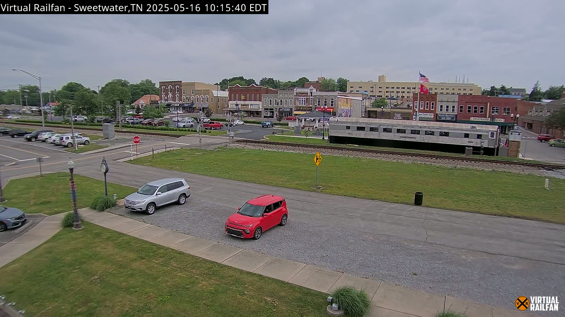 A small town's downtown area under overcast skies, with several cars parked near a railway track where a long train car is situated.