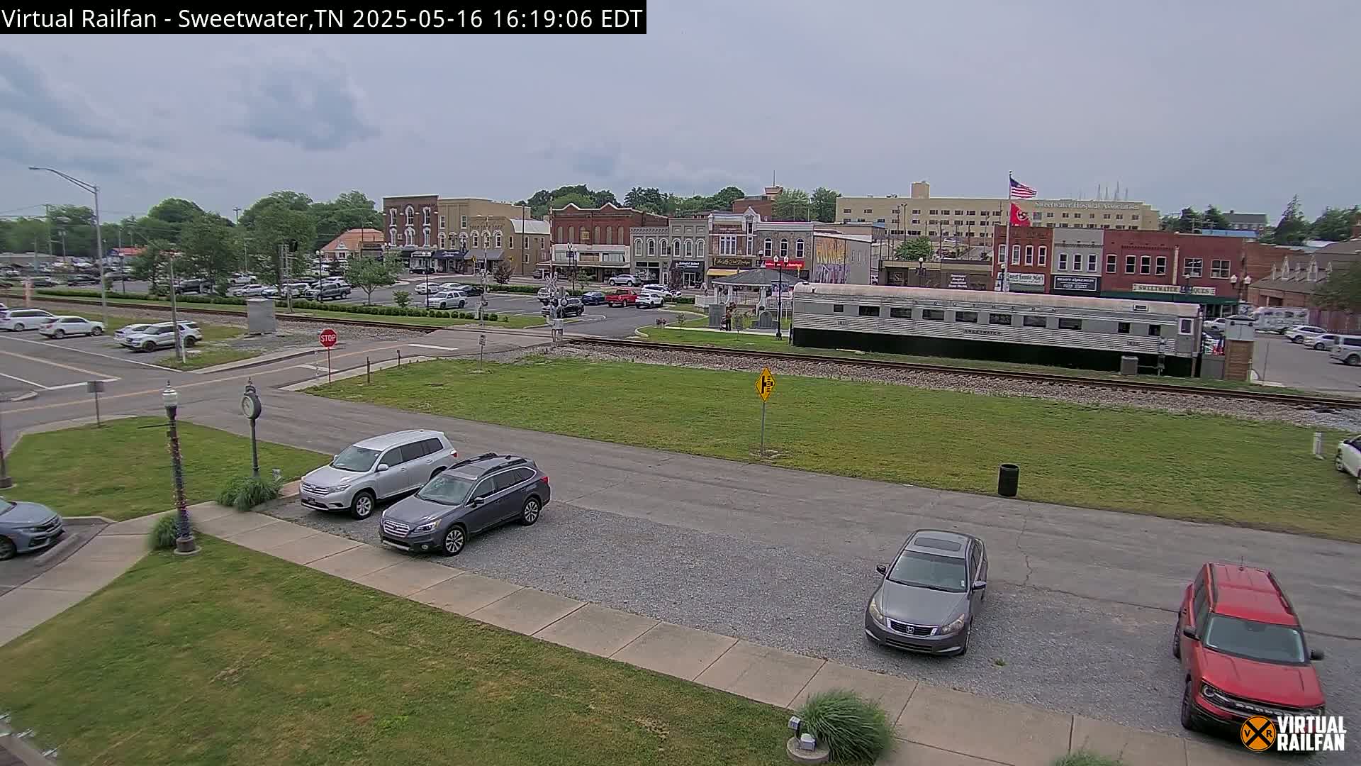A mostly cloudy day shows a small town square with parked cars and a silver passenger car on a railway track in front of brick buildings.