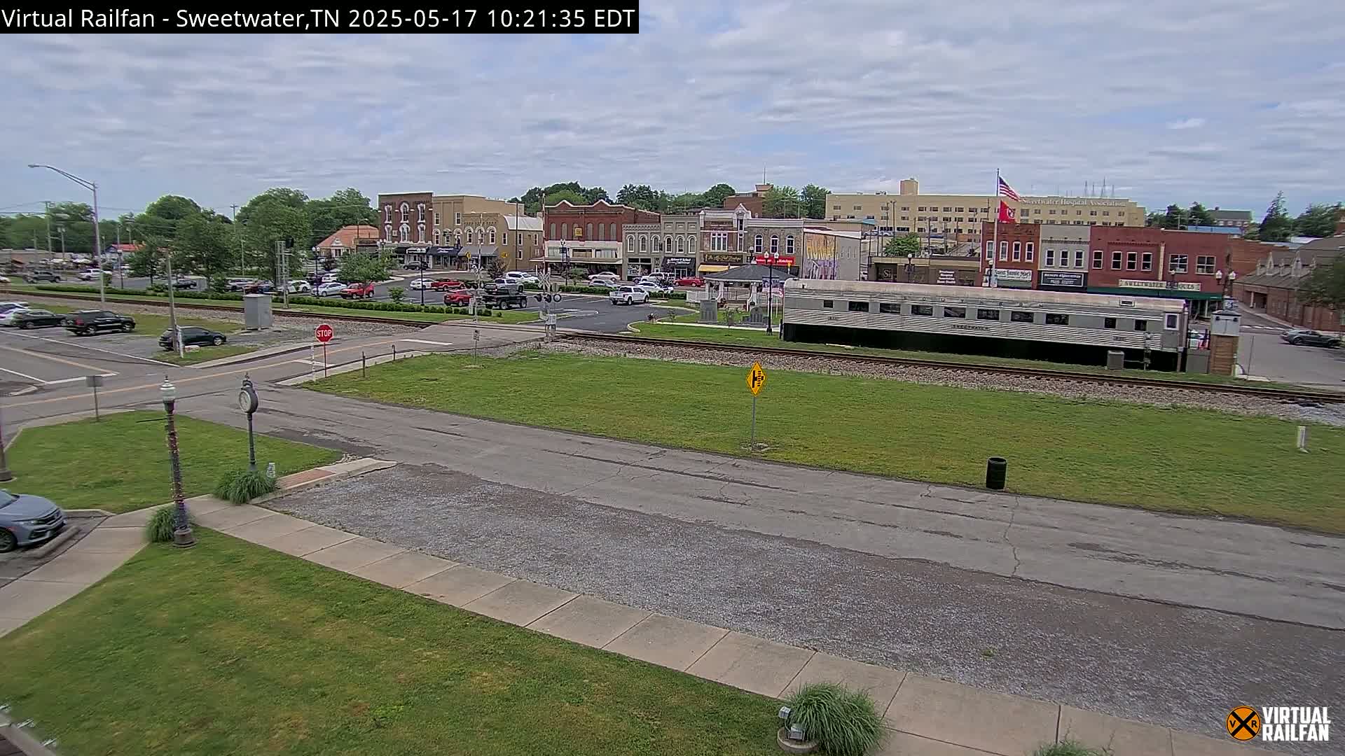 A partly cloudy day shows a town square with several buildings, cars, and a railway car parked on the tracks.