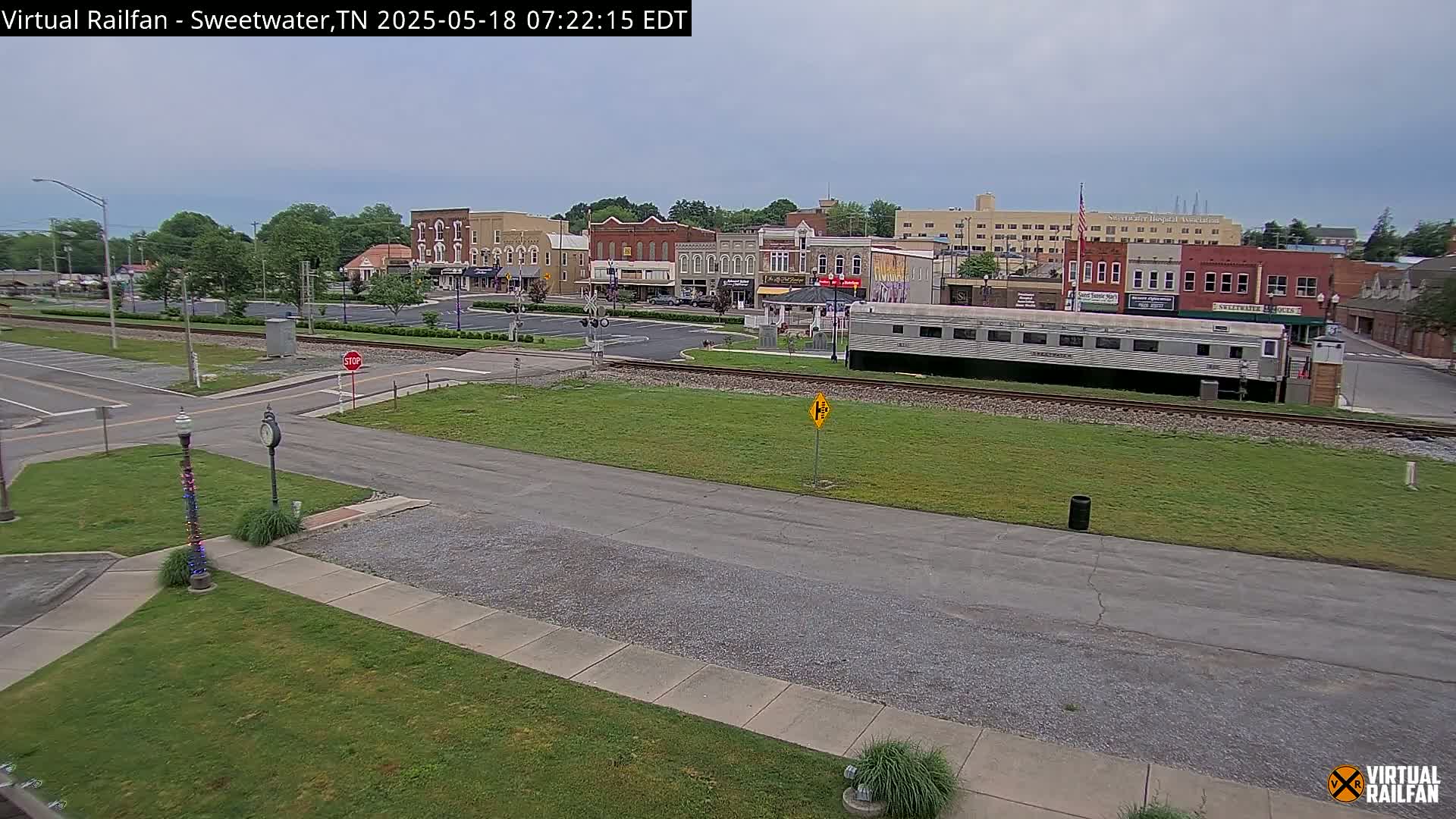 A mostly cloudy day reveals a small town square with several brick buildings, a train car parked on nearby tracks, and a mostly empty parking lot.