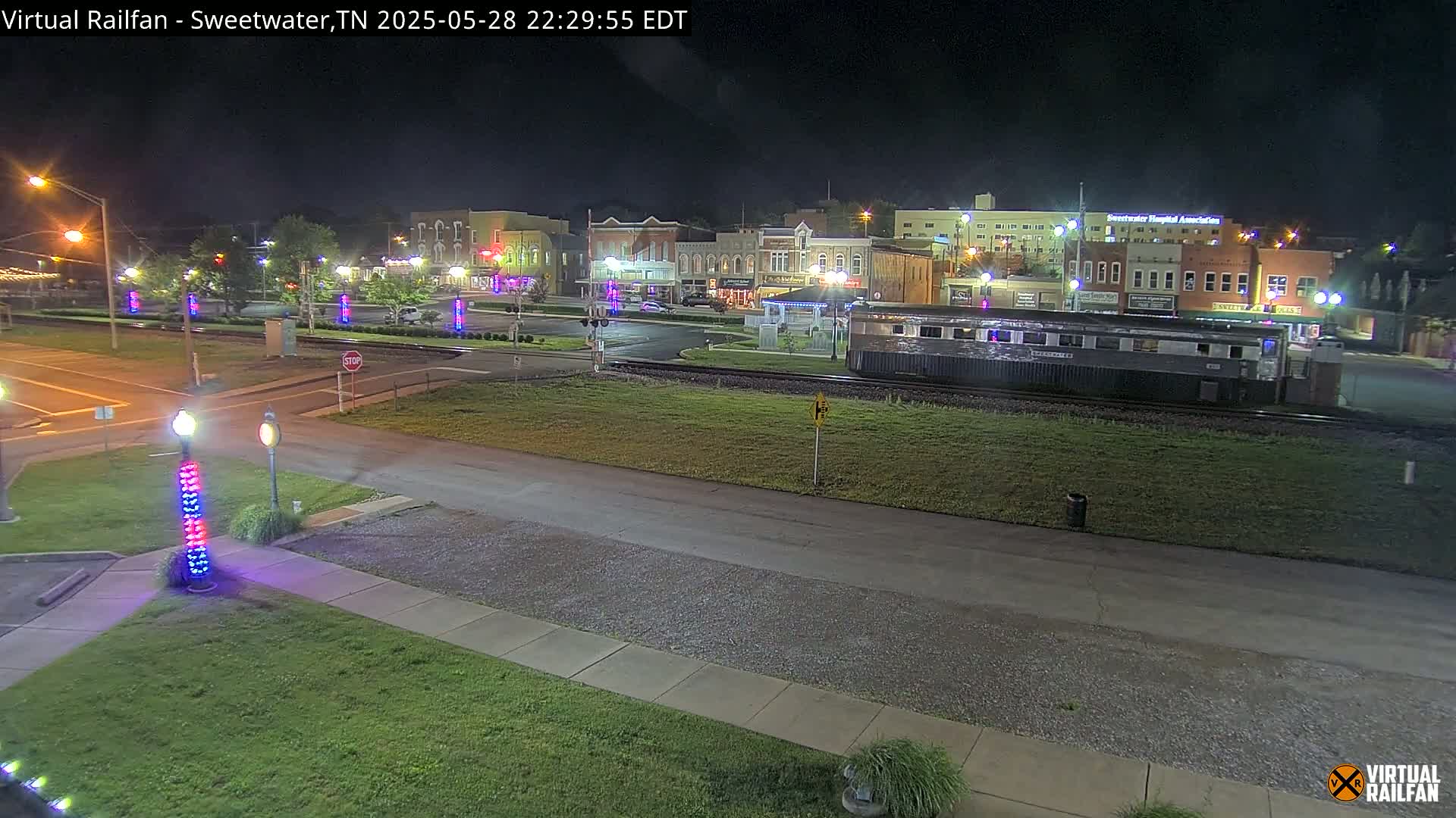 A nighttime view of a small town's downtown area, featuring a railroad car parked on tracks in the foreground, several buildings illuminated, and light posts with decorative lighting.