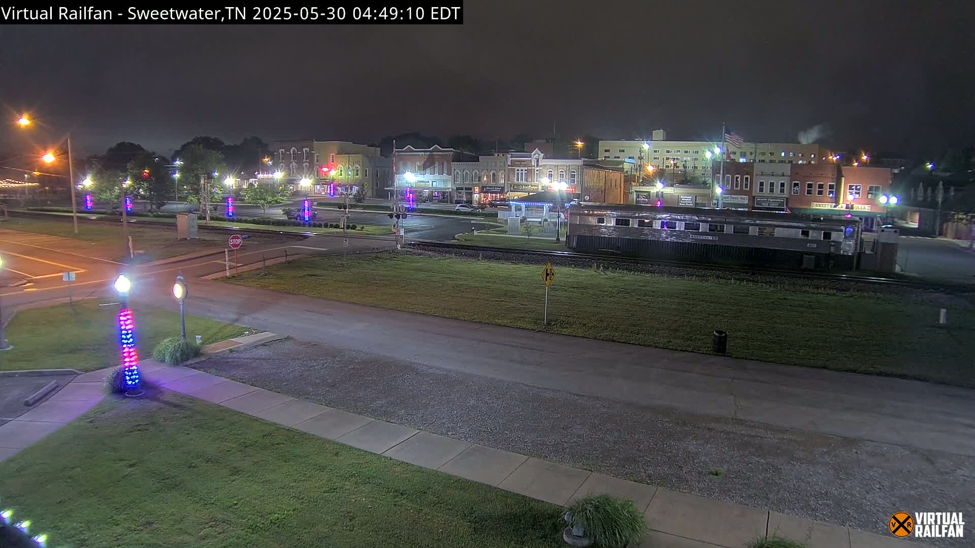 A nighttime view of a town square with several buildings, a railway car, and decorative light posts, under a dark sky.