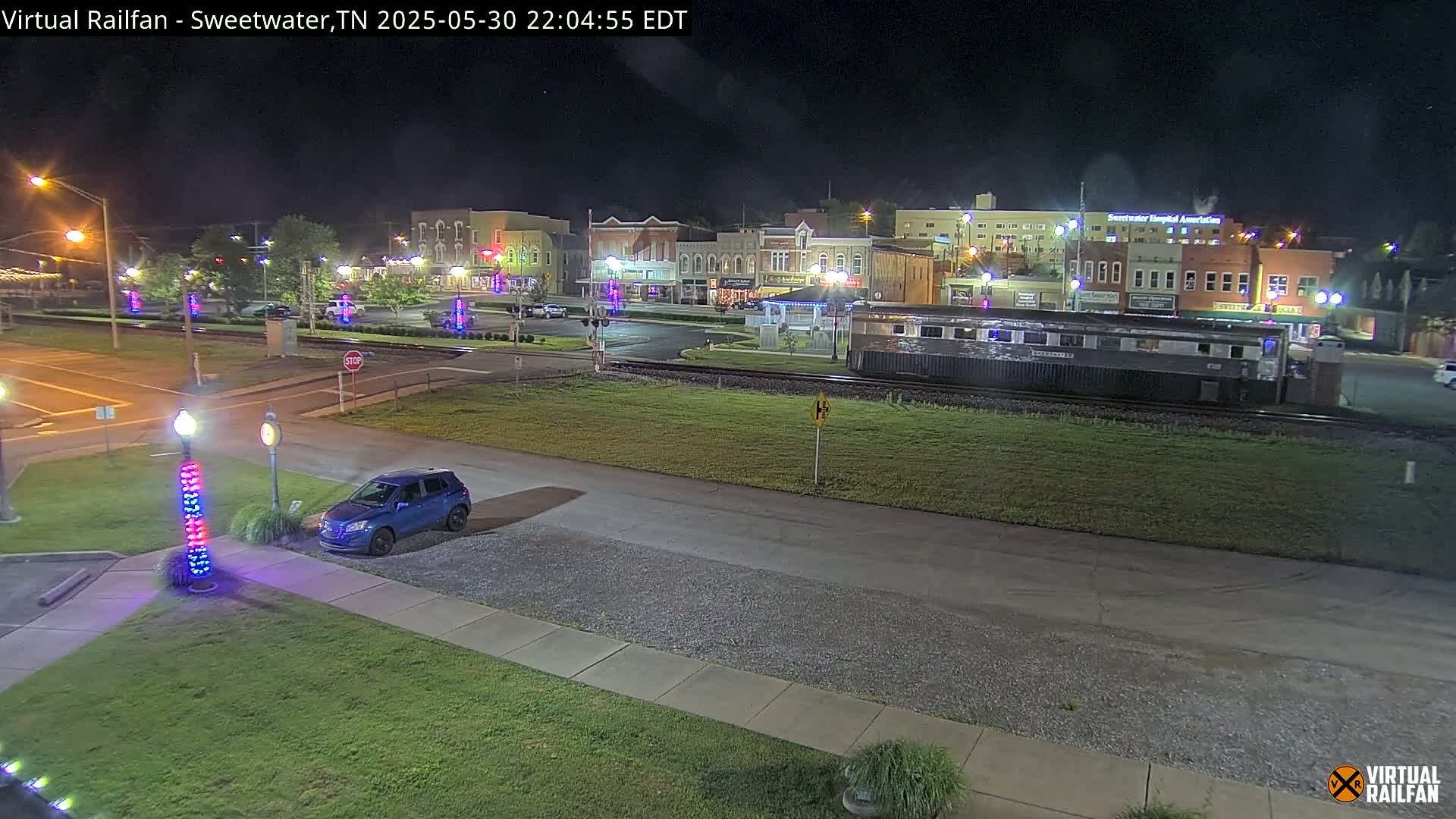 A small town's nighttime streetscape is shown, featuring a parked blue car near a rail line with a train car, and a row of buildings illuminated with decorative lights.