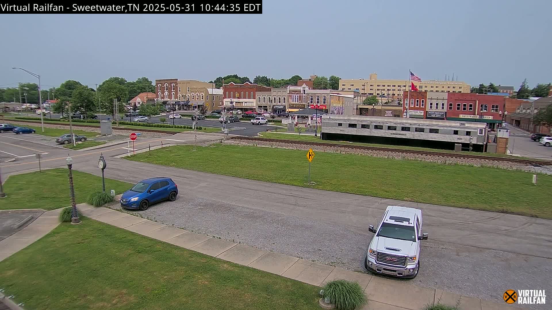 A mostly sunny view of a small town's downtown area with several cars parked in a lot, and two cars parked on the street near a railway line with a train car.