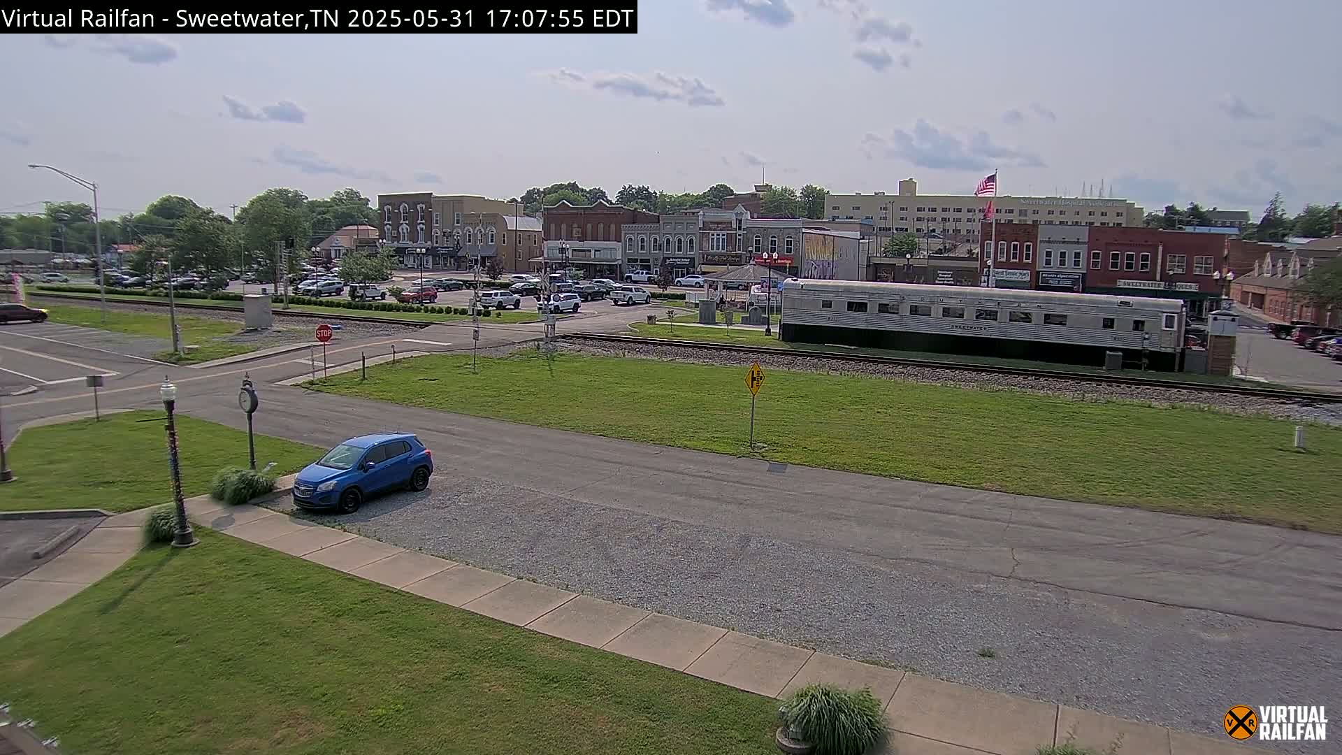 A blue car is parked in a lot overlooking a town square with a train car and several brick buildings under a partly cloudy sky.