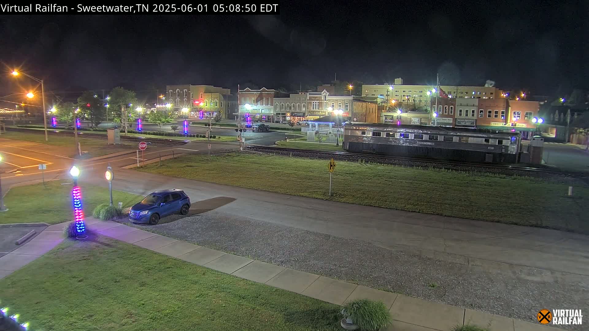 A nighttime view of a small town square with a single blue car parked near a railway line where a train car is sitting, with illuminated light posts lining the streets.