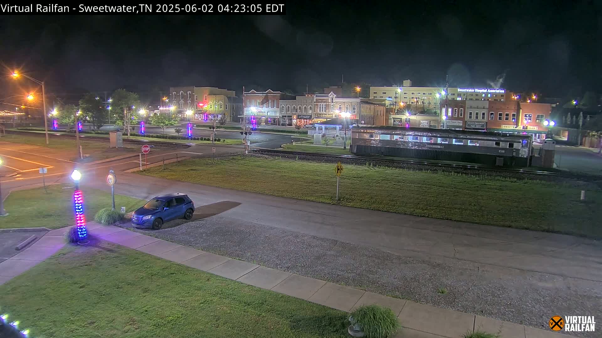 A nighttime view of a town square with a train car parked nearby, illuminated by streetlights, with a single blue SUV parked in a lot.
