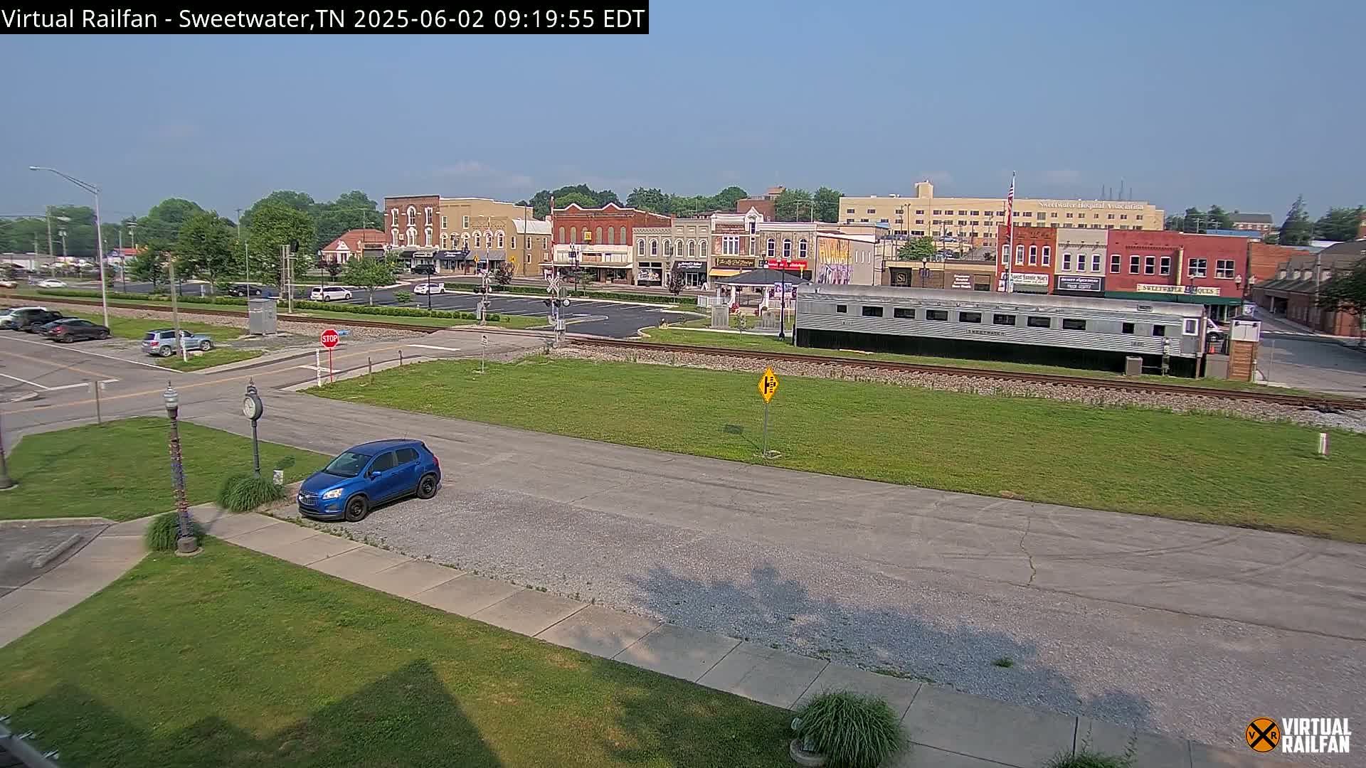 A blue car is parked in a lot across from a railroad track with a long silver passenger car next to it, and a town square with brick buildings is visible in the background under a clear sky.