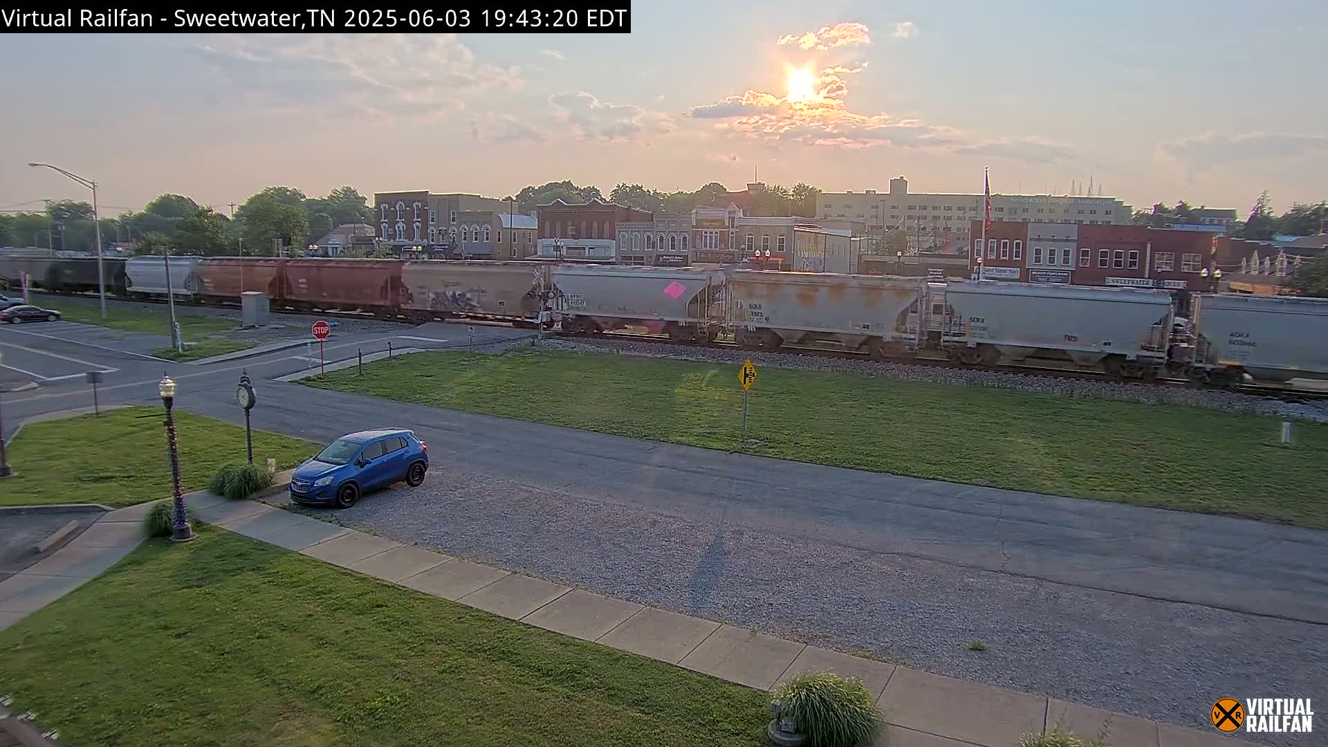 A long freight train passes through a small town under a partly cloudy sunset sky, with a blue SUV parked nearby.