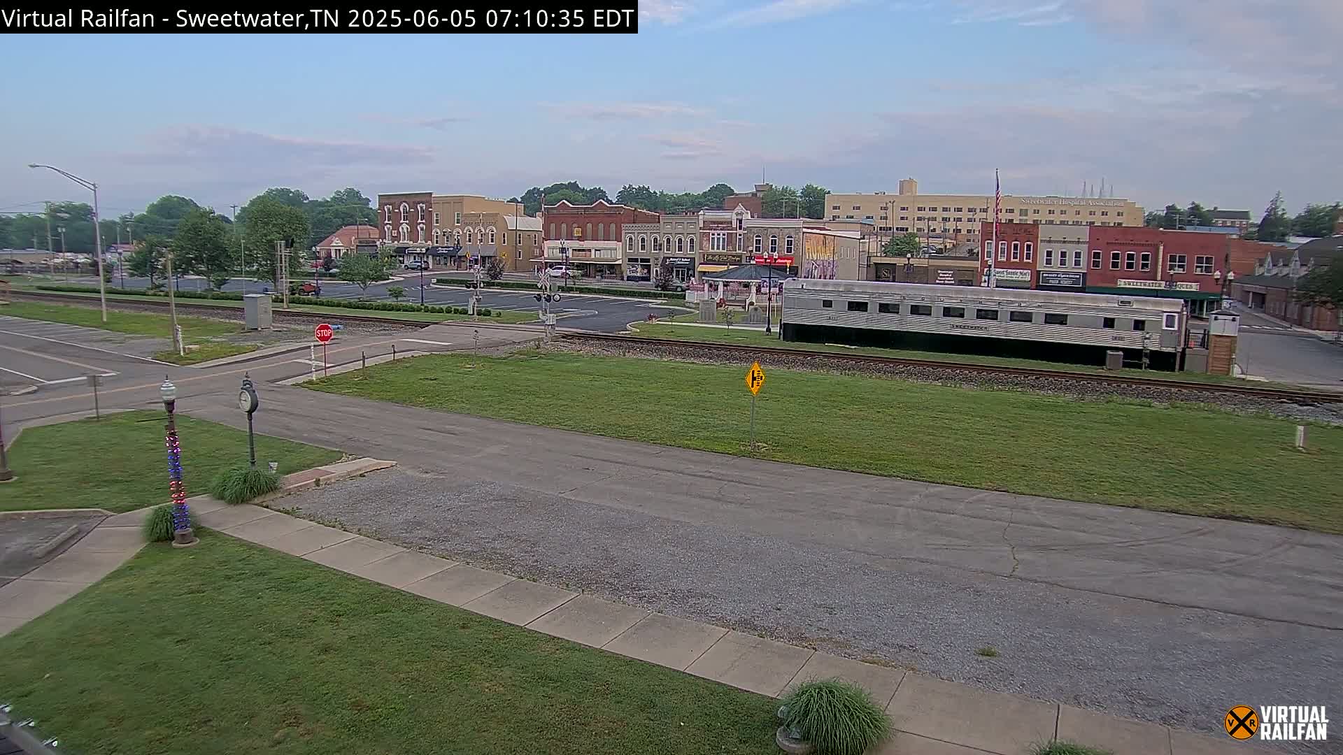 A mostly sunny view of a small town's downtown area, featuring several brick buildings, a railway line with a long silver passenger car, and a grassy area.