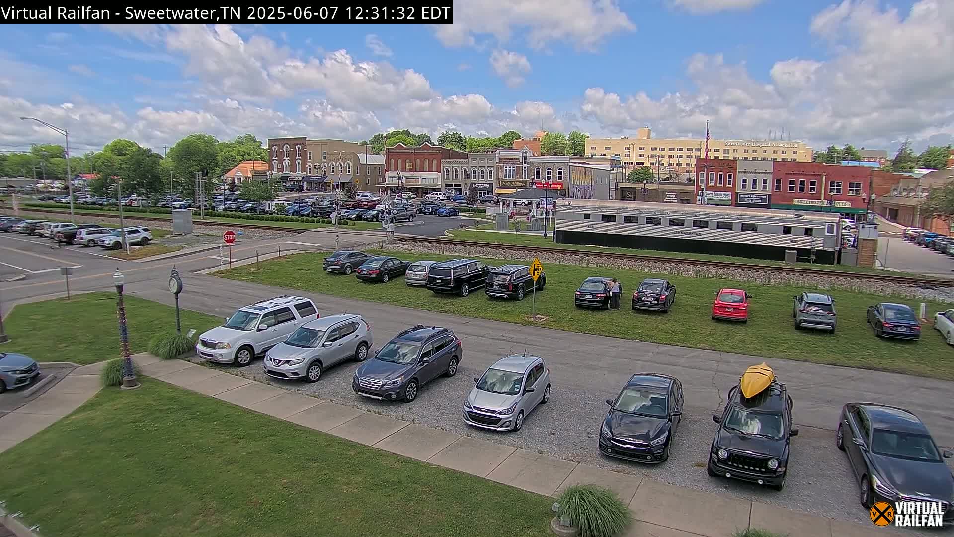 A partly cloudy day reveals a town square with several parked cars, a train car, and multiple brick buildings.