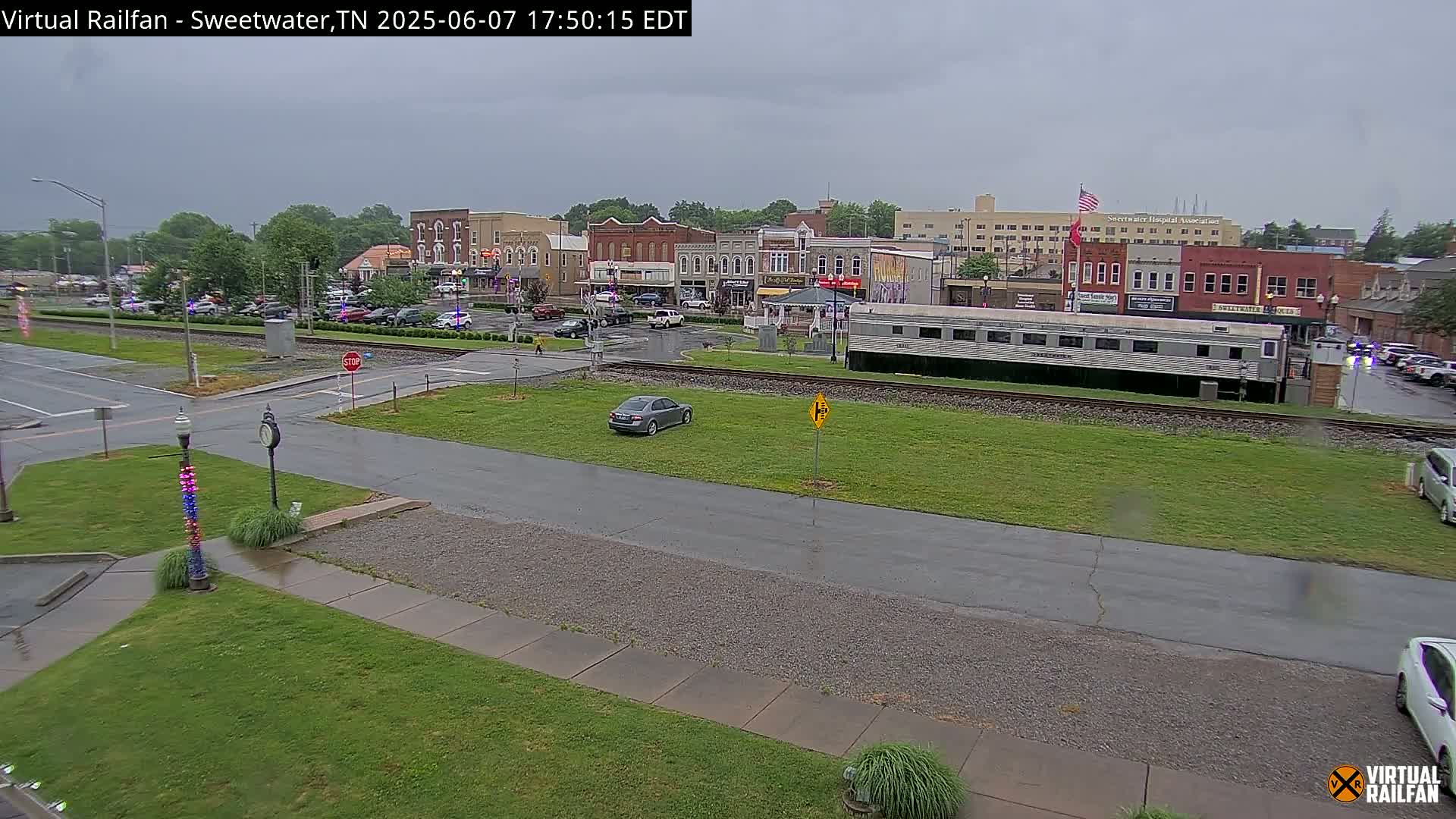 A rain-slicked town square with several buildings, a parked train car, and a few cars is visible in an overcast setting.