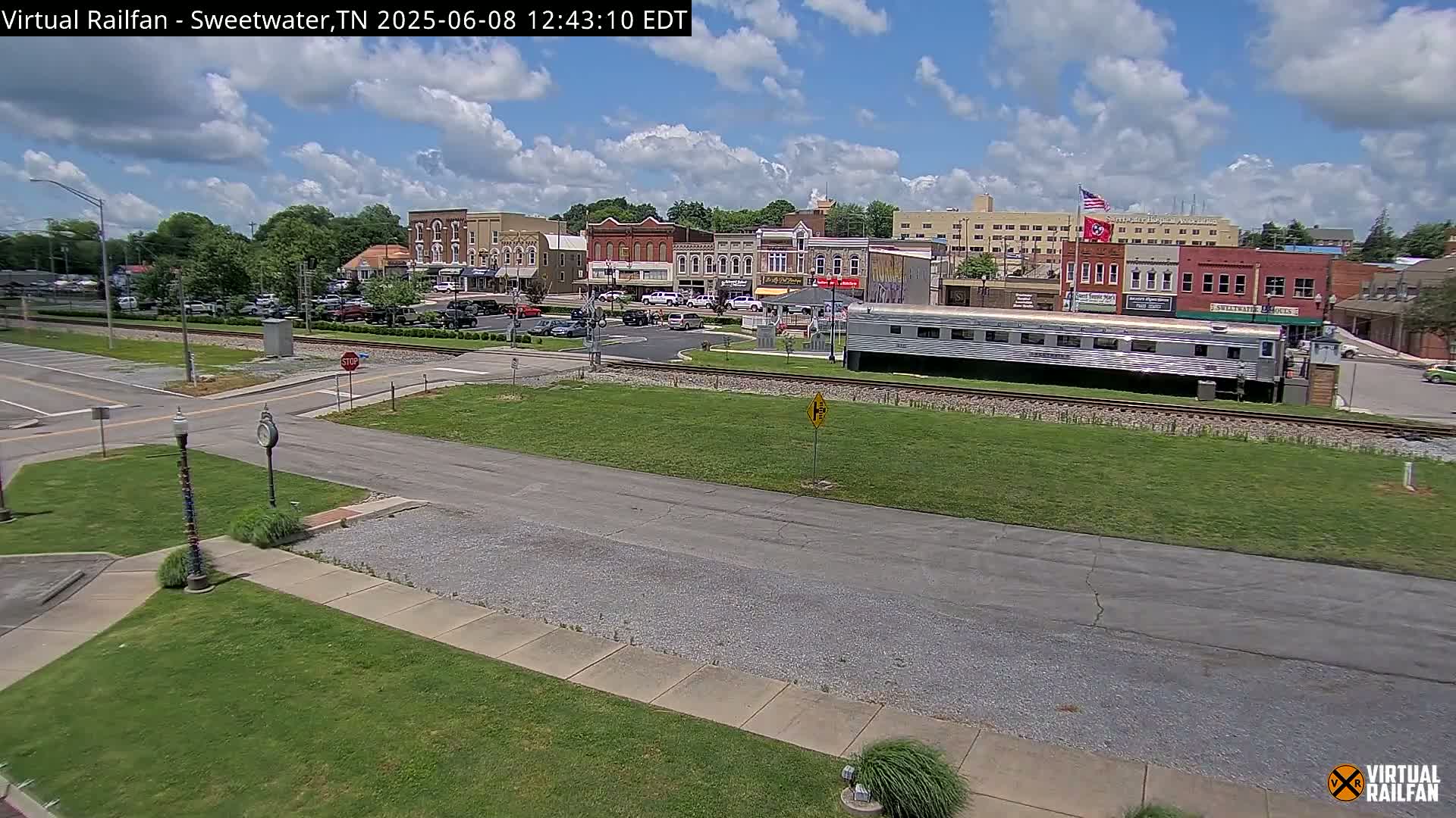 A partly cloudy day reveals a town square with several buildings, a railroad track with a long silver train car parked alongside, and a grassy area.