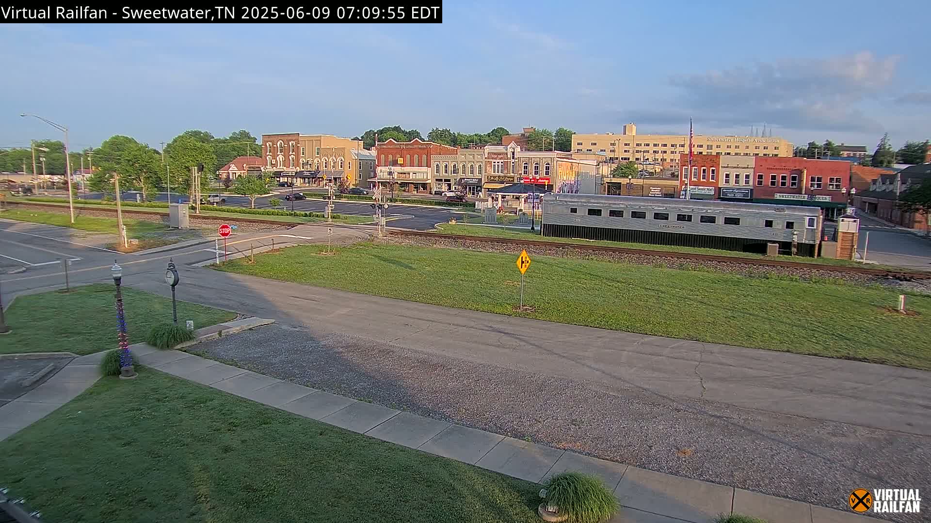 A mostly sunny view of a small town's downtown area, featuring several brick buildings, a parked railway car, and a mostly empty town square.