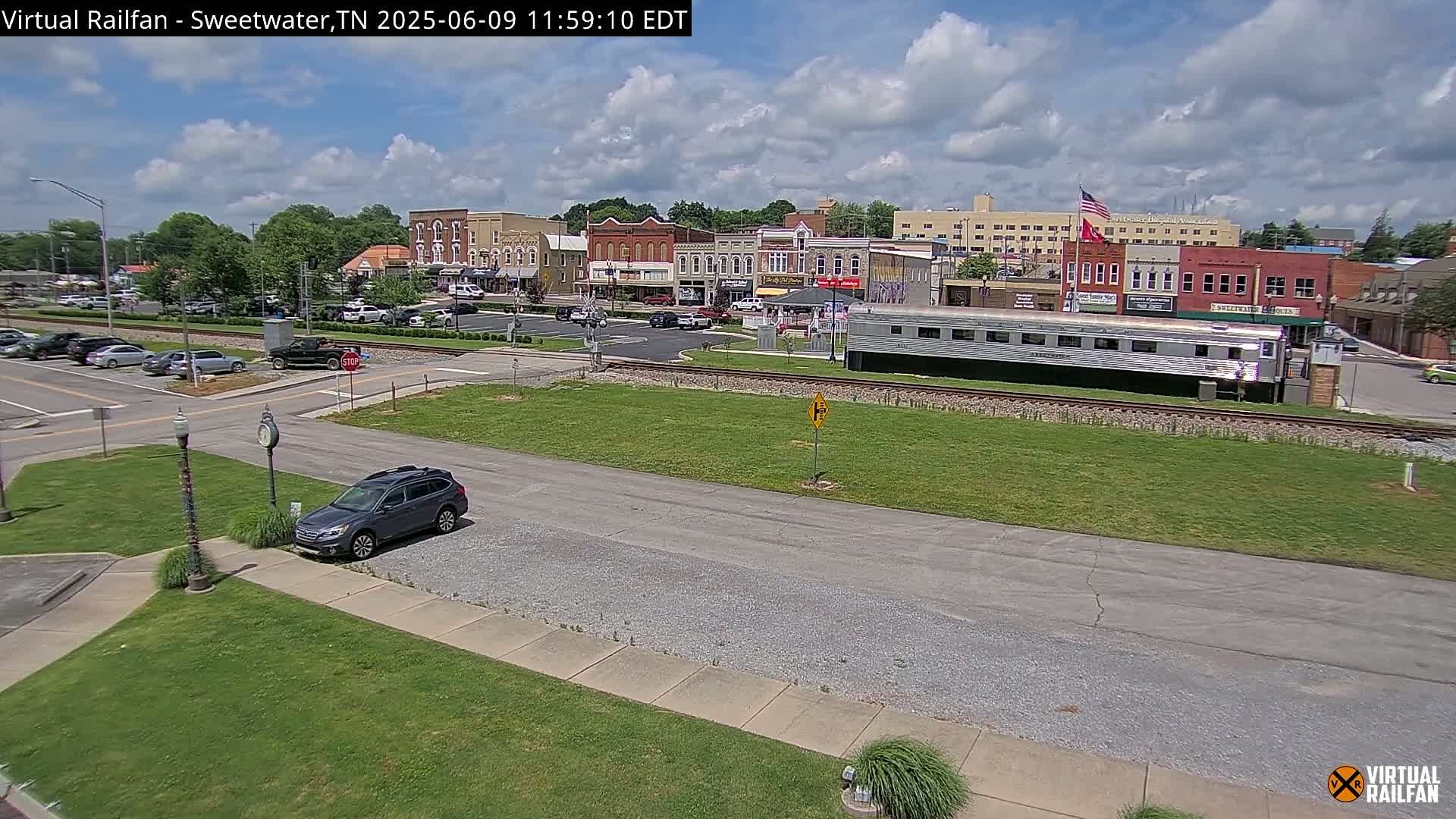 A mostly sunny day reveals a small town square with parked cars, a long silver train car, and several brick buildings, all situated beside train tracks.