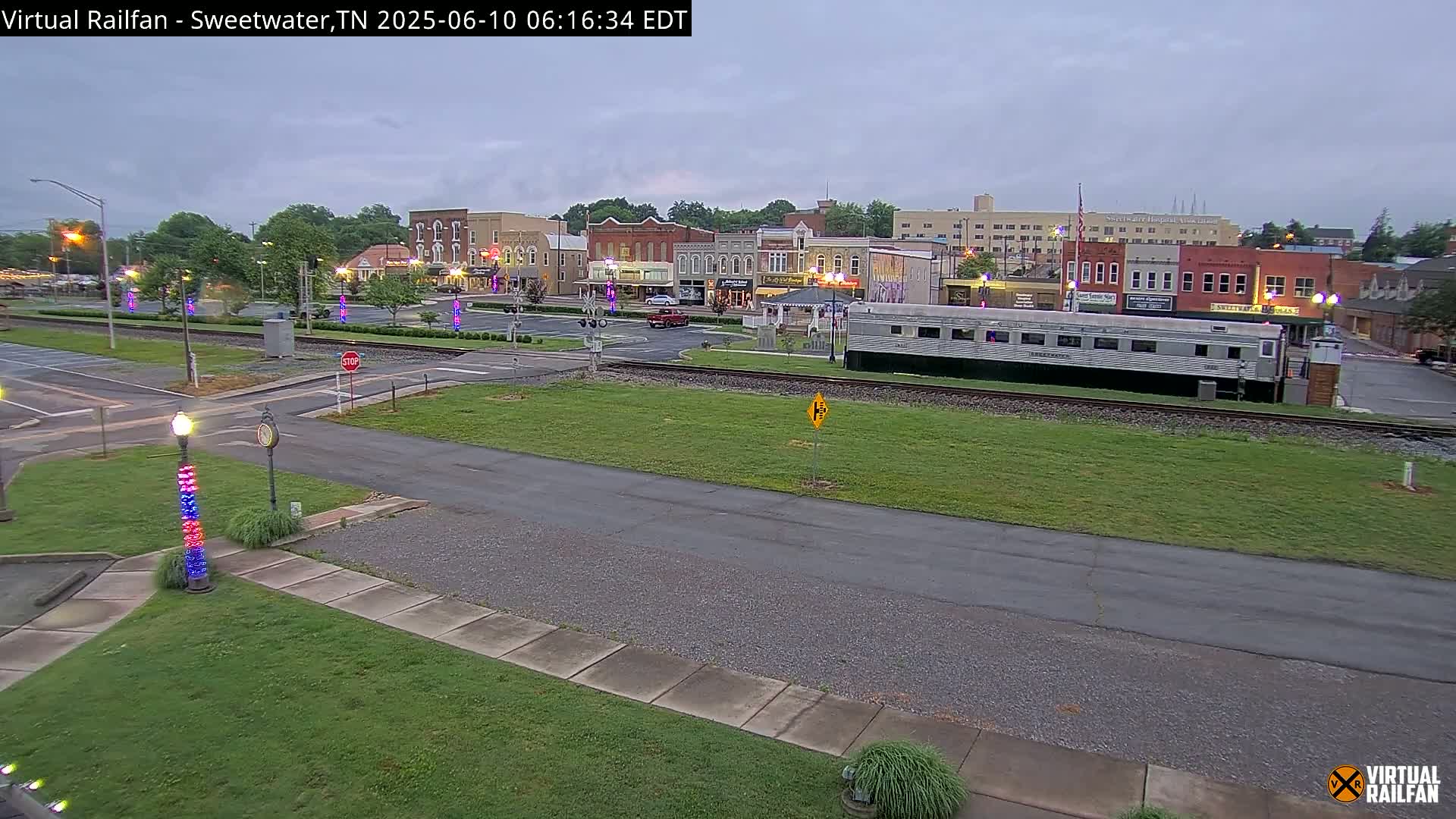 A view of a town square with several buildings, a railway track with a passenger car parked nearby, and overcast skies.