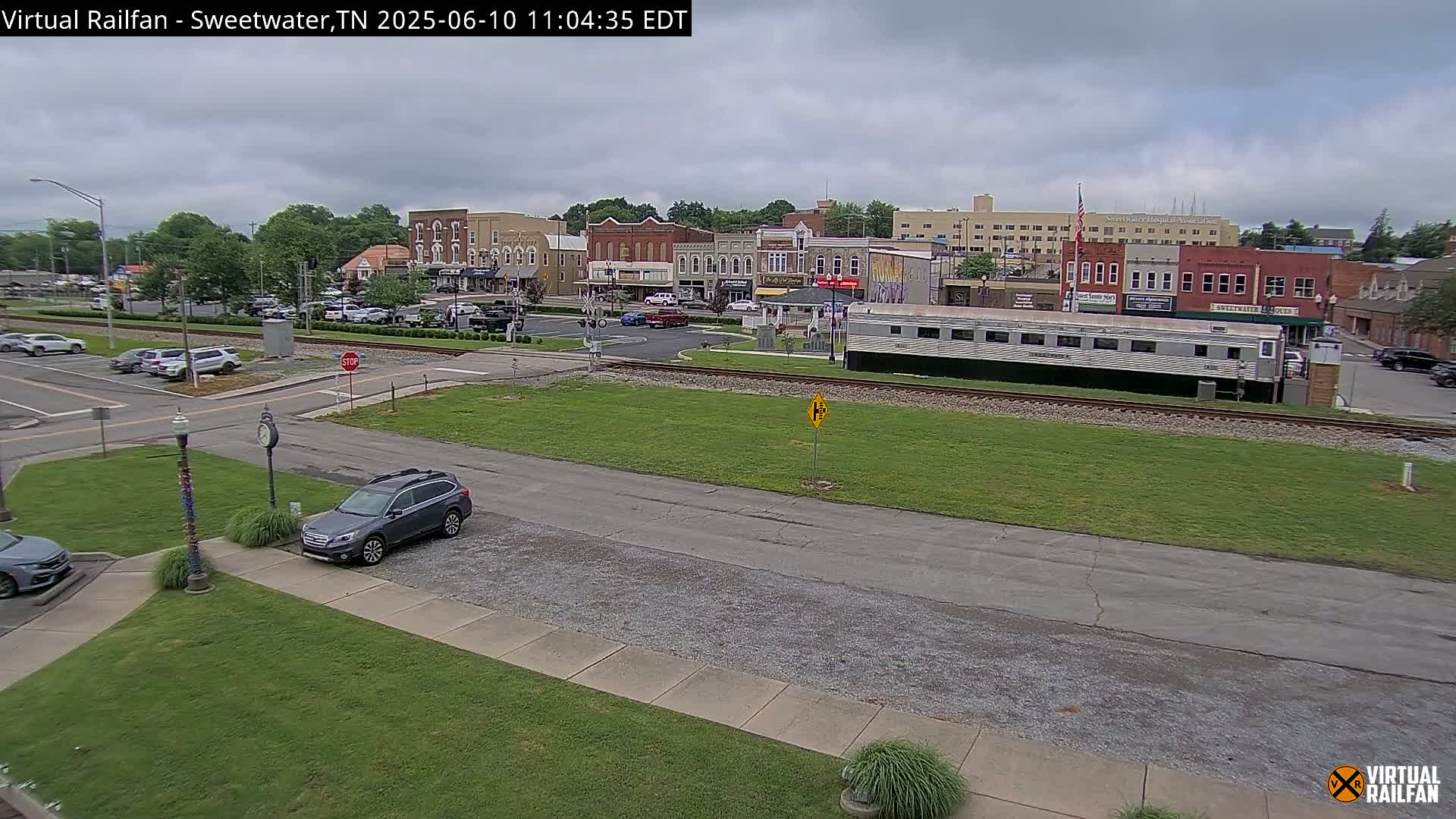 A mostly cloudy day shows a small town's downtown area with several brick buildings, a railway track with a passenger car parked alongside, and several cars parked in lots and driving on the streets.