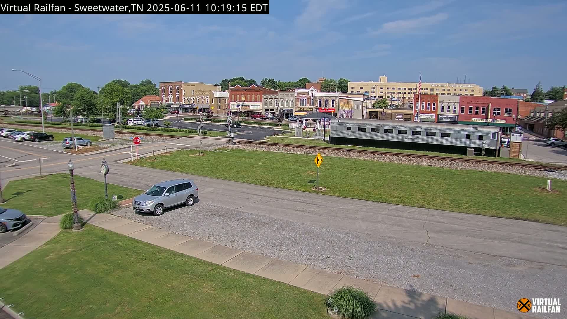 A silver SUV is parked near a railroad track with a long silver passenger car, in front of a small town's downtown area on a sunny day.